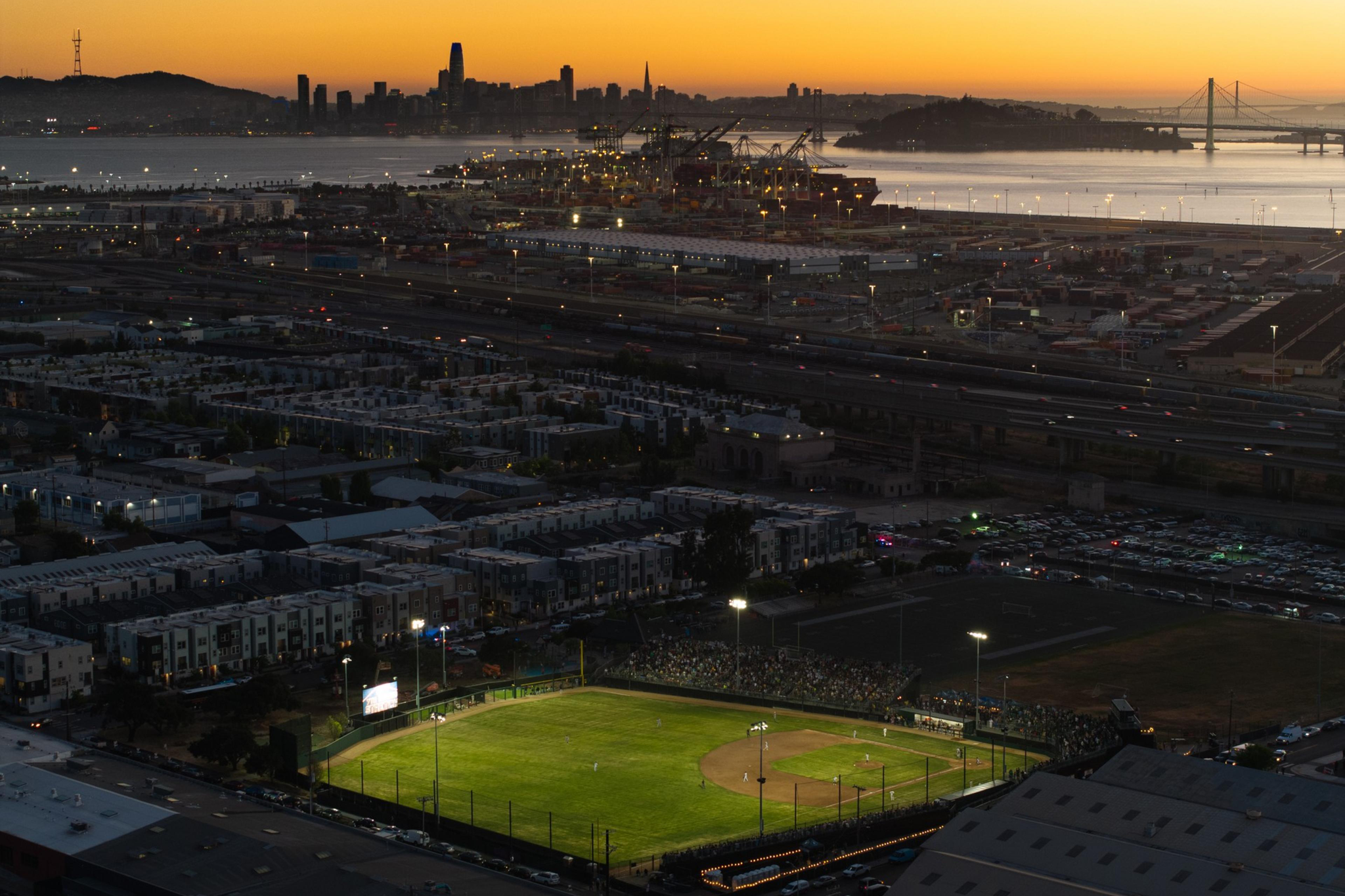 The image shows a baseball field lit up for a night game, surrounded by a cityscape, with a sunset over a water body and distant bridge and city skyline.