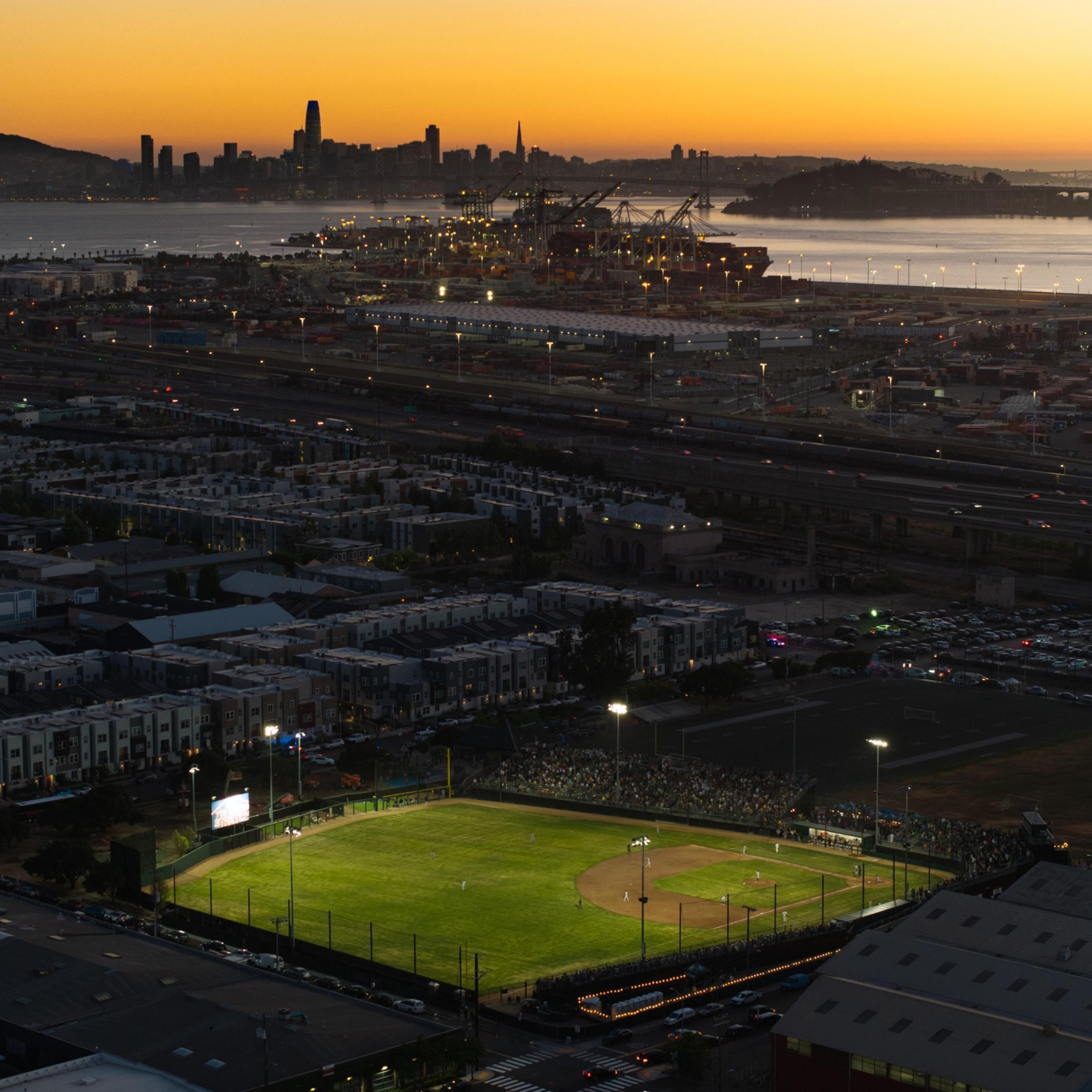 The image shows a baseball field lit up for a night game, surrounded by a cityscape, with a sunset over a water body and distant bridge and city skyline.