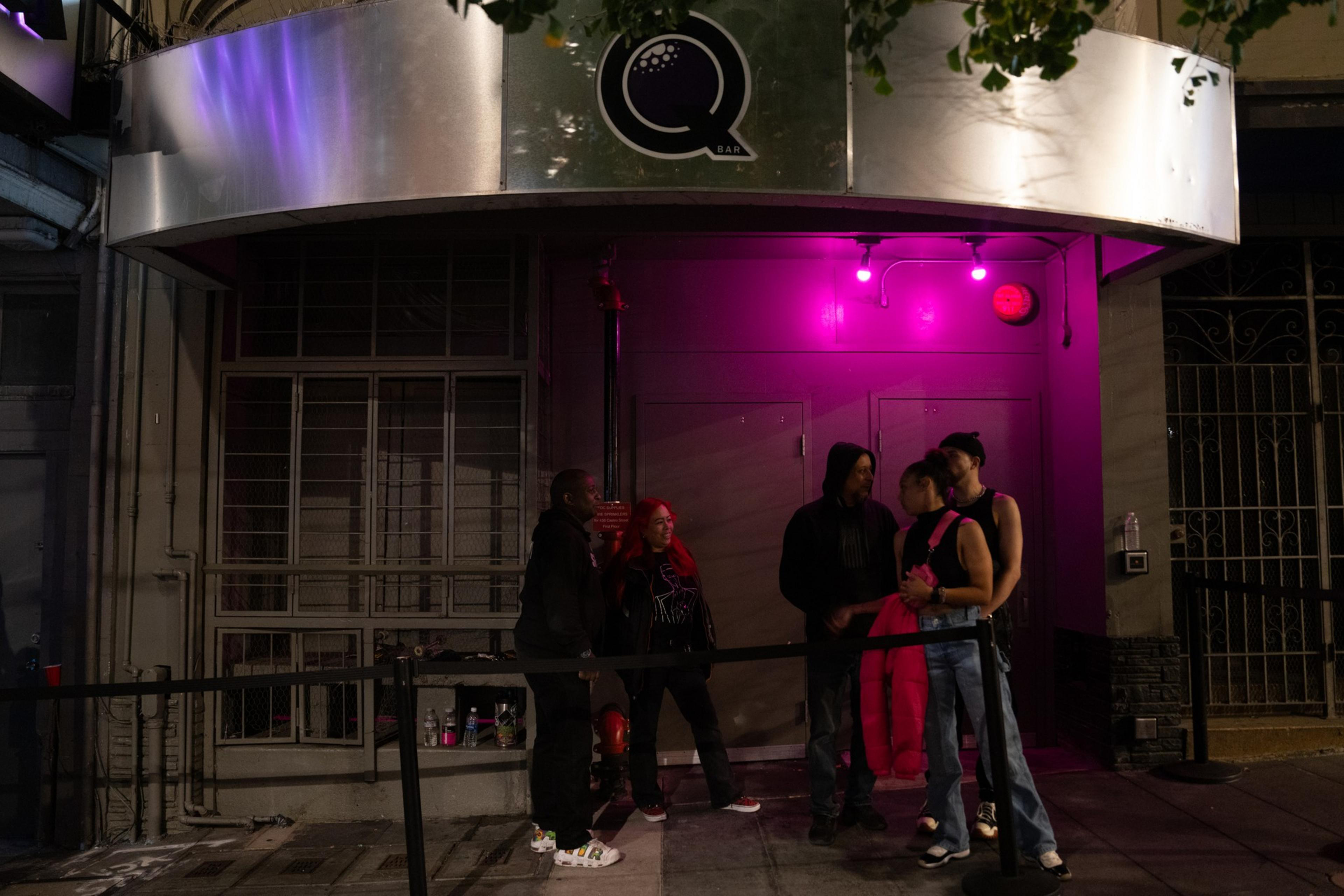 A group of people stands outside a bar with a &quot;Q&quot; logo. The scene is dimly lit by pink lights, with some people chatting and others standing silently.