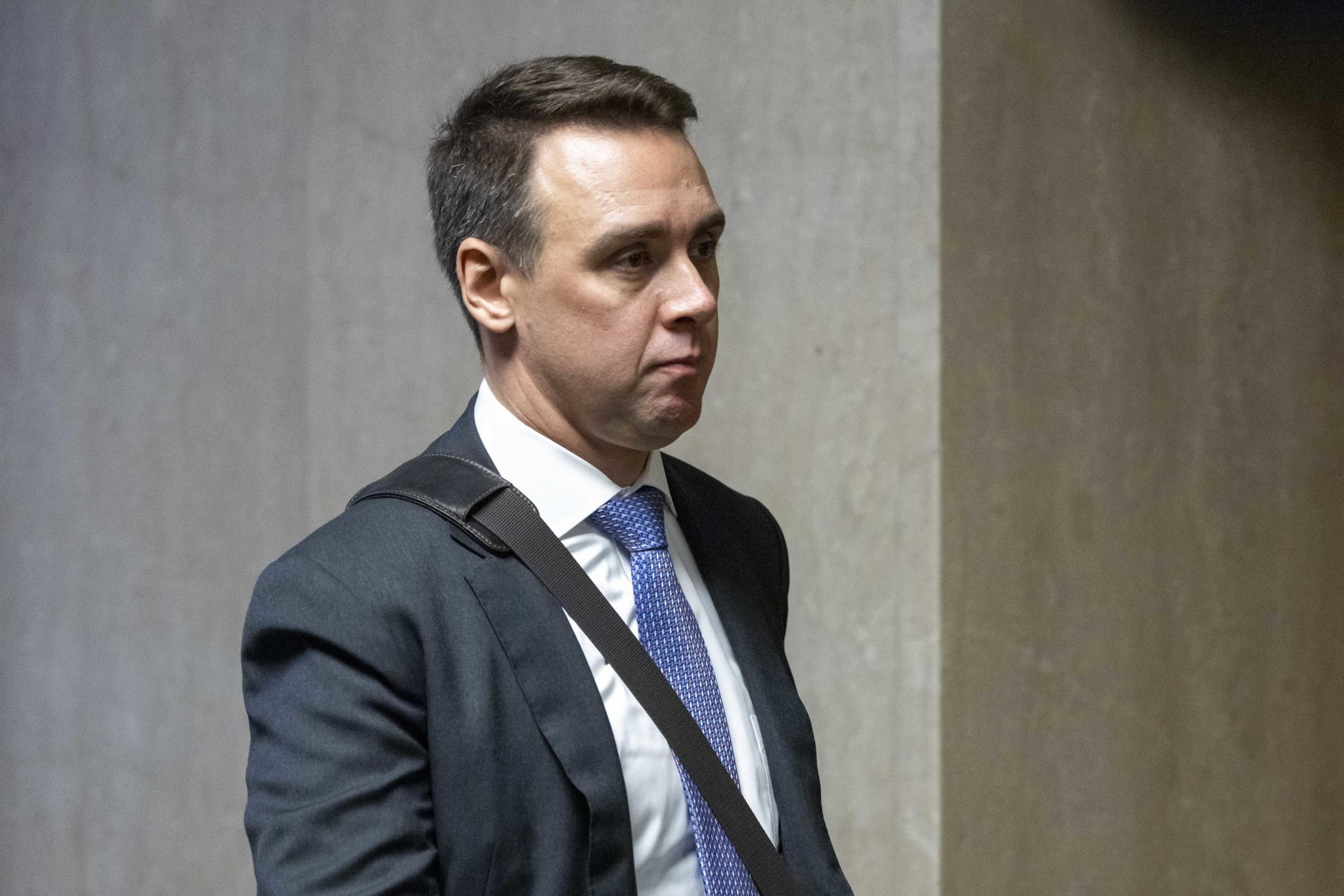 A man in a suit with a blue tie and a strap across his shoulder is standing against a neutral background, looking slightly to the side.