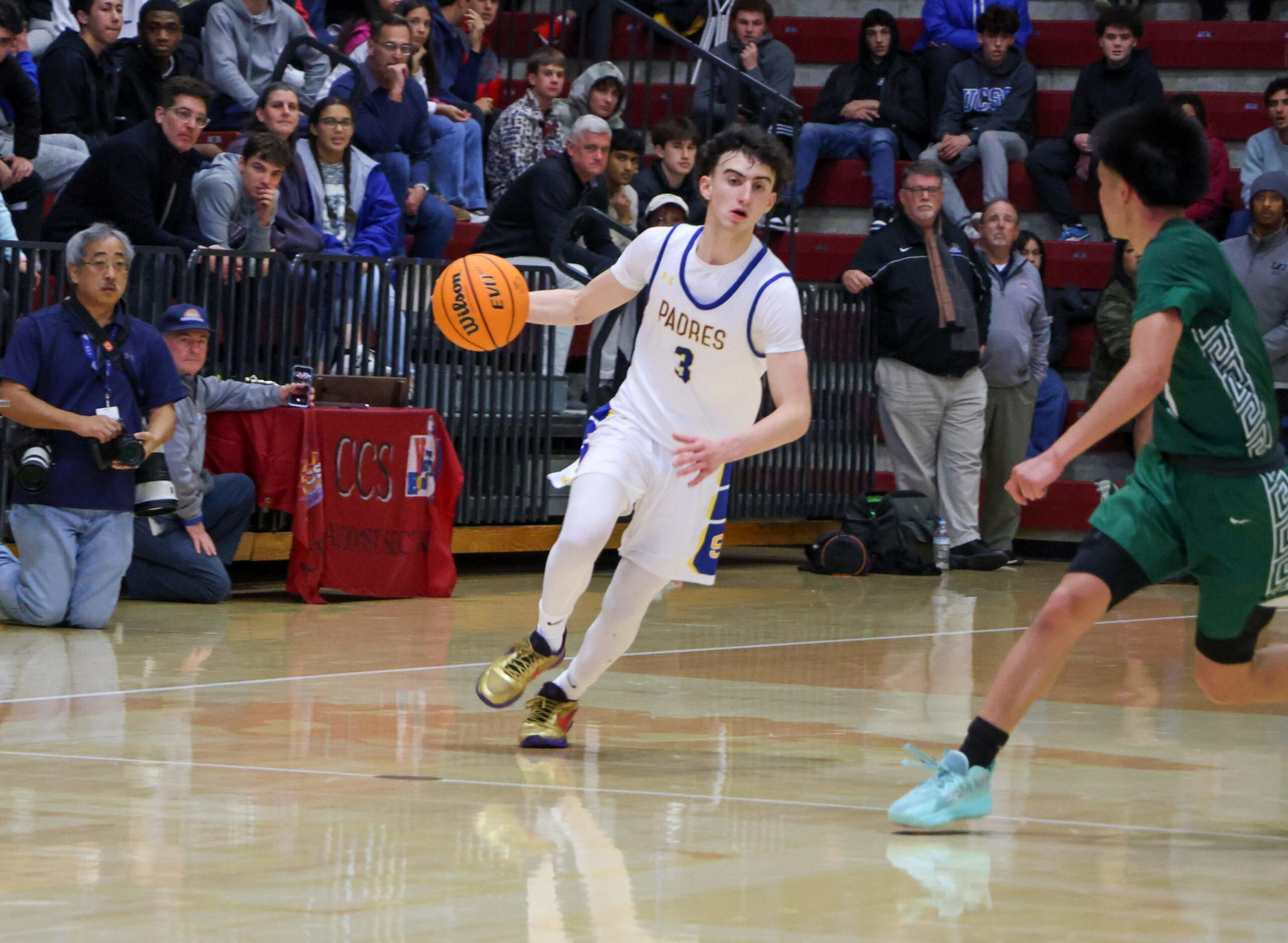 A basketball player in a white "Padres" uniform dribbles the ball while guarded by a player in green, with spectators watching from the bleachers.