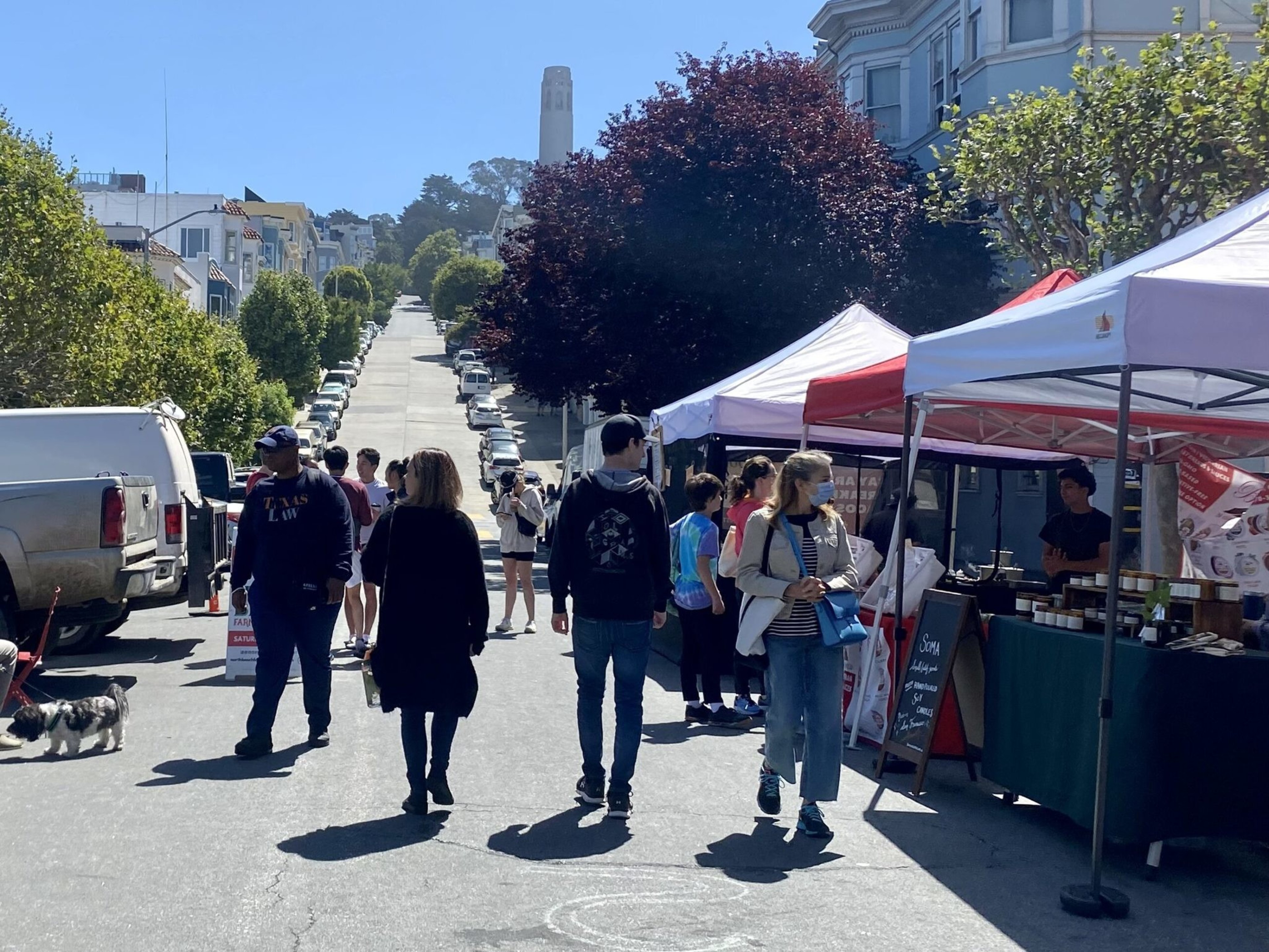 People walk along a street lined with market tents on one side, with cars parked on both sides and a tall white tower visible in the distance.