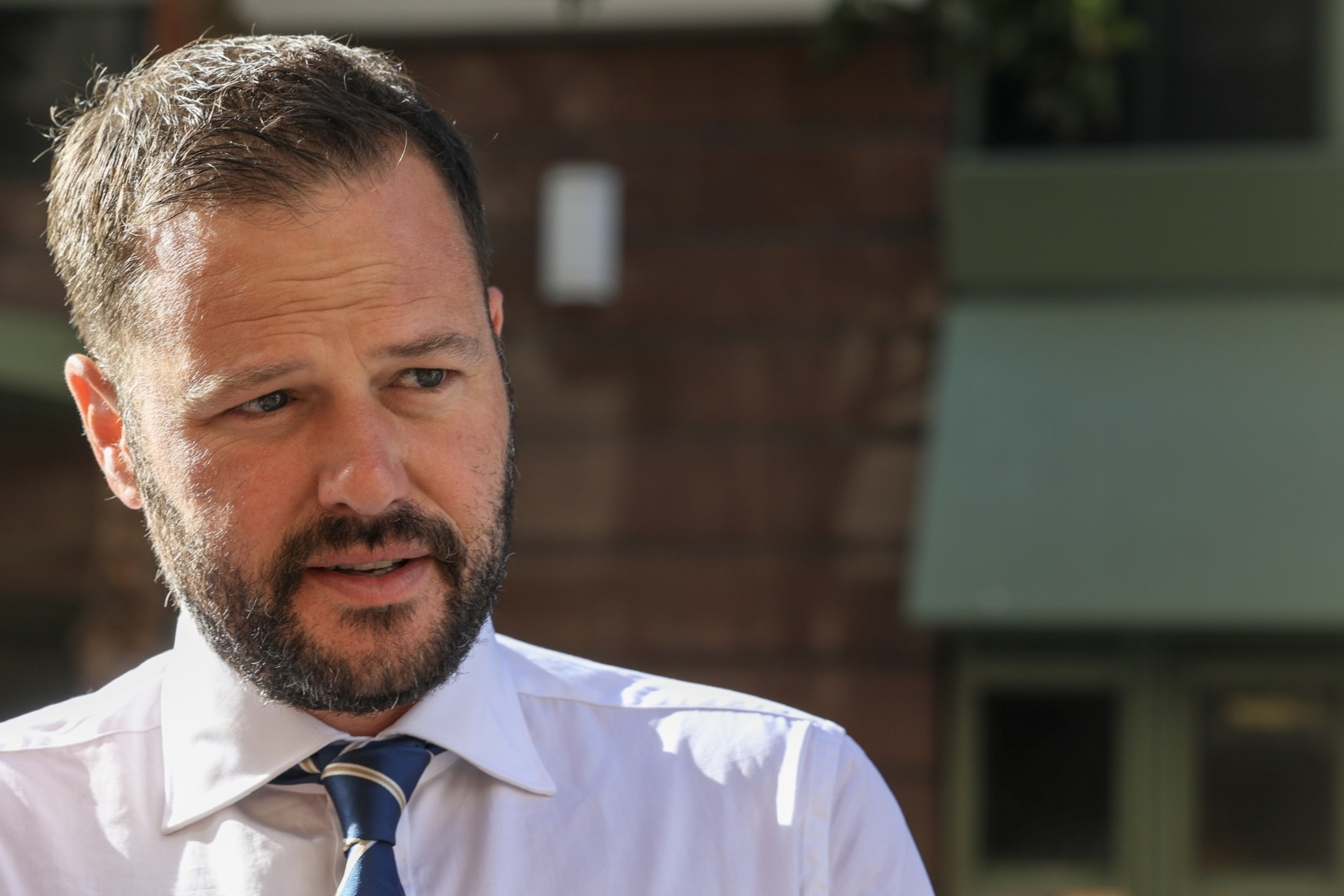 A man with short brown hair and a beard is wearing a white dress shirt and a striped tie. He is outdoors, set against a blurred background of a building facade.