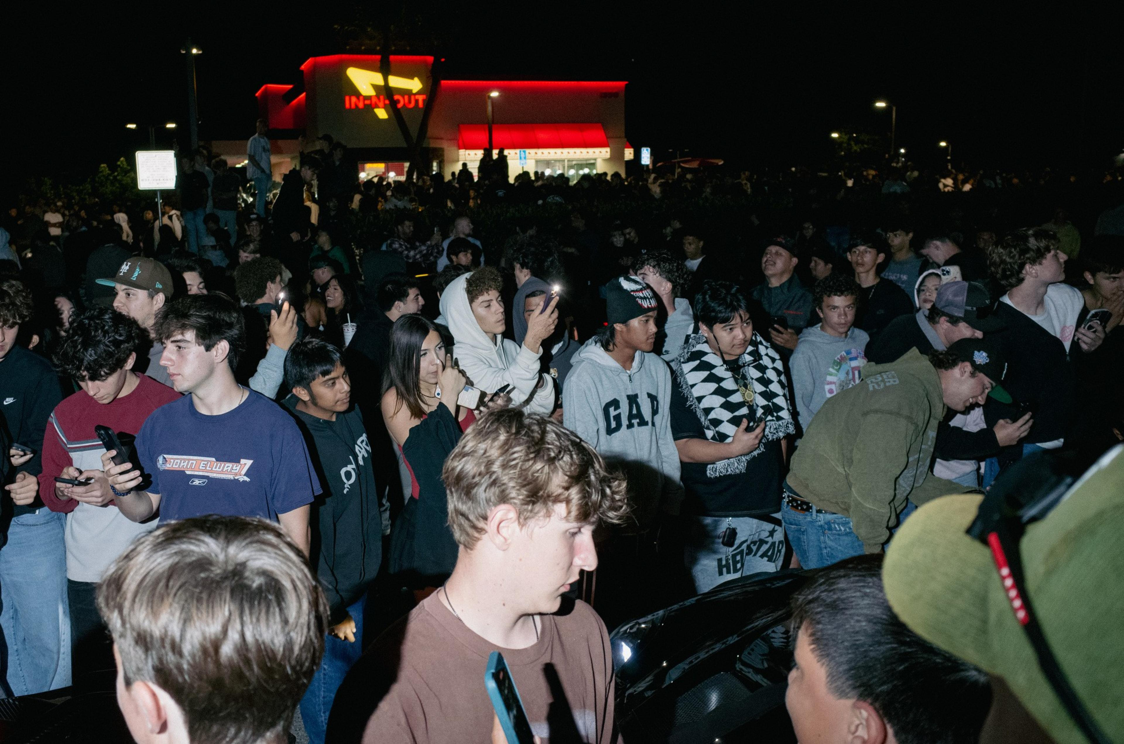 A large crowd of mostly young people gathers outside an In-N-Out restaurant at night, many holding phones and looking in various directions.