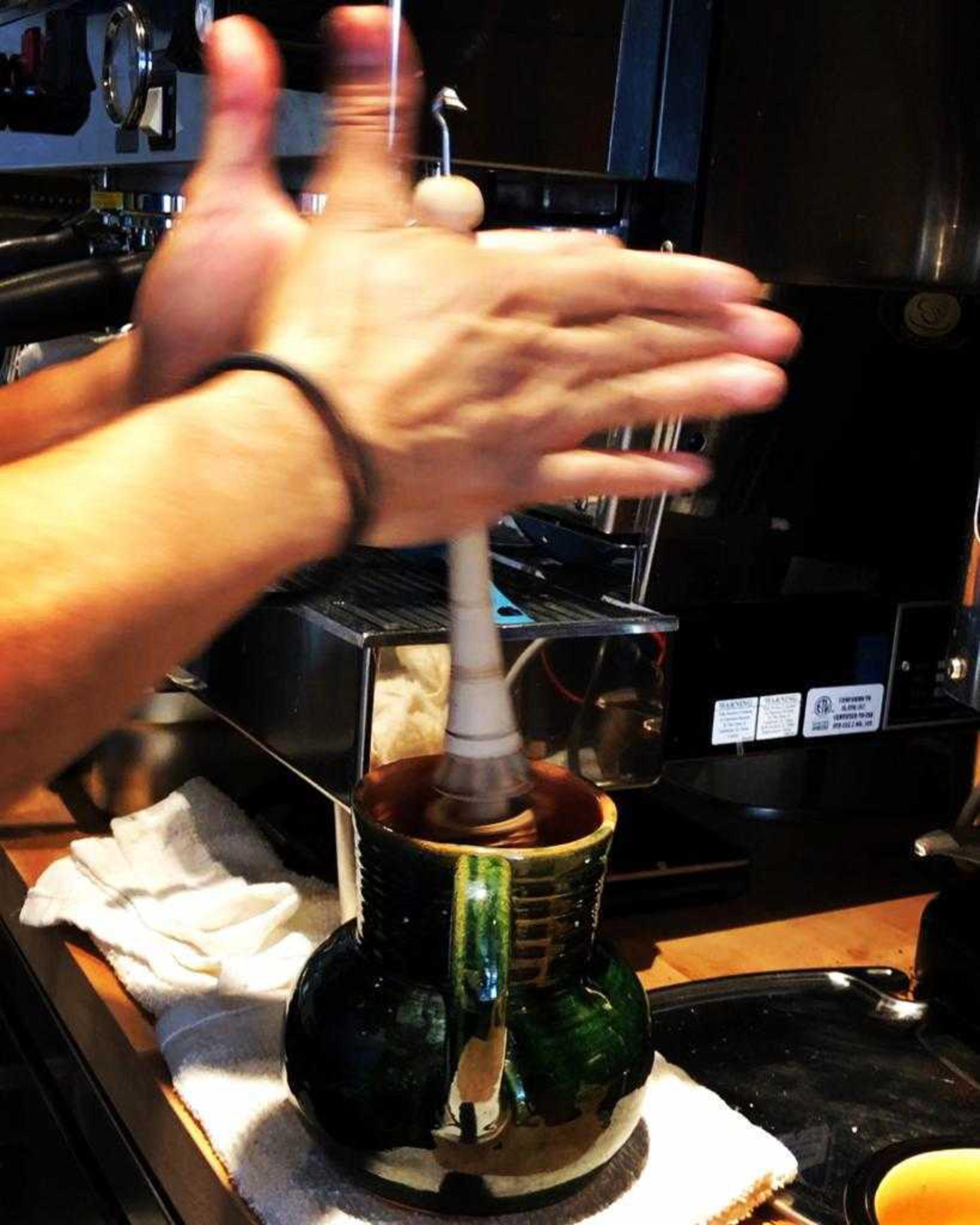 A pair of hands is frothing milk in a green ceramic pitcher on a coffee machine counter with a white towel beneath.
