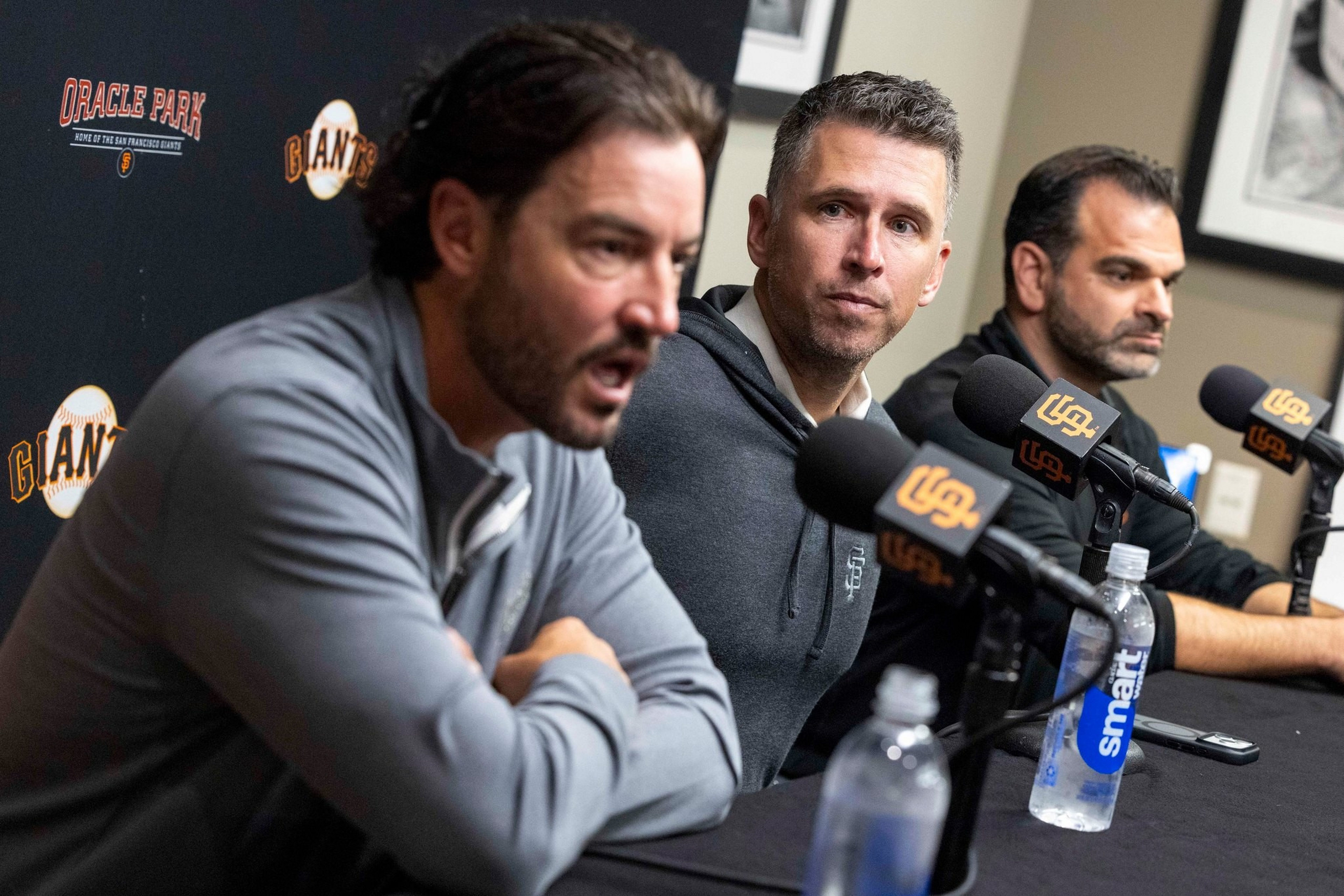 Three men sit at a table speaking into microphones with "USA" logos in front of a backdrop reading "Oracle Park" and "Giants."