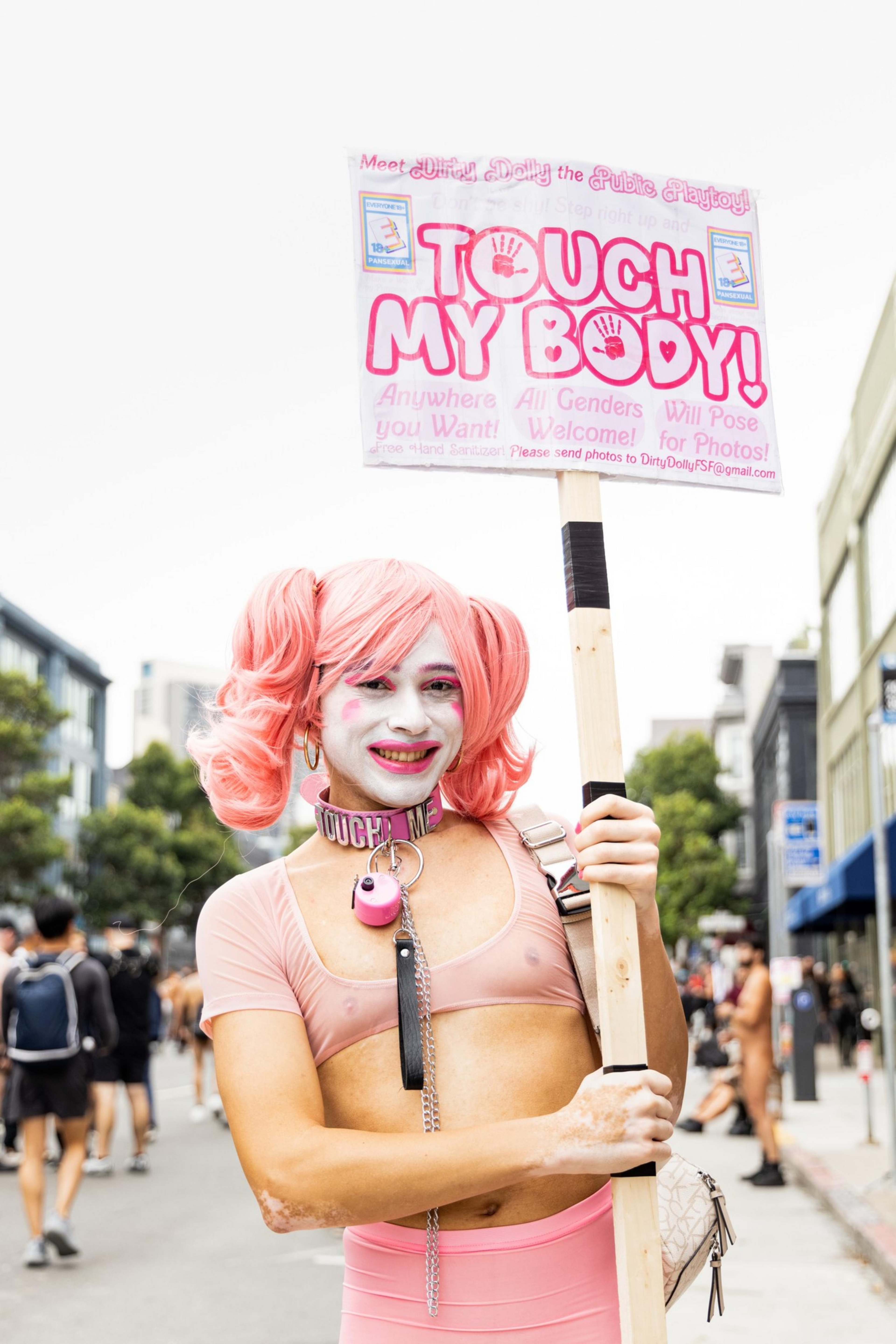 Person with pink hair in pigtails, white face paint, and pink outfit holds a sign saying “TOUCH MY BODY!” in a busy outdoor urban setting.
