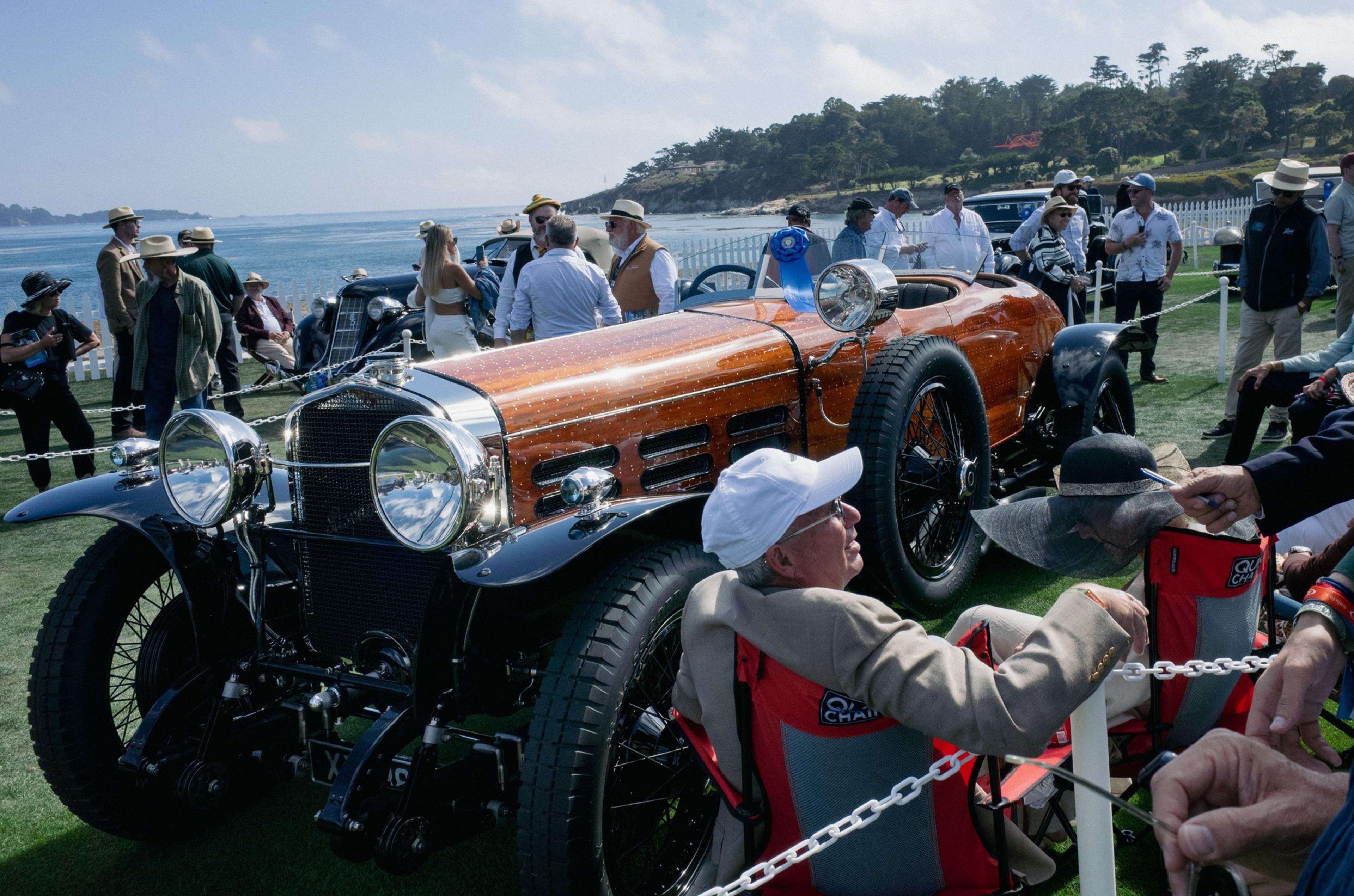 A vintage orange and black car is displayed on grass by the sea, surrounded by people admiring it, with a man in a white cap sitting nearby.