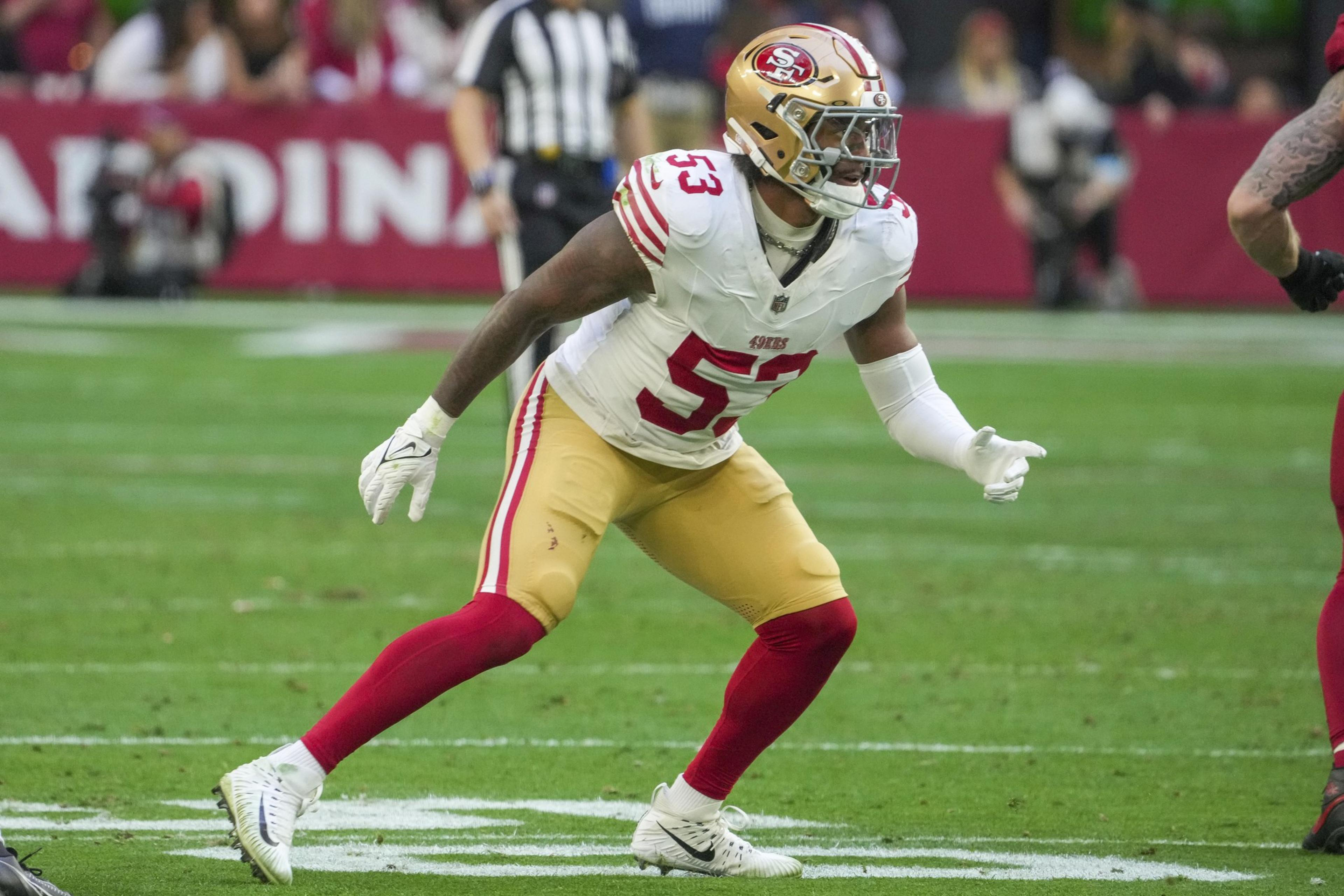 A football player wearing a white 49ers jersey and gold helmet is in an action stance on a grass field. He's wearing red leggings and white cleats.
