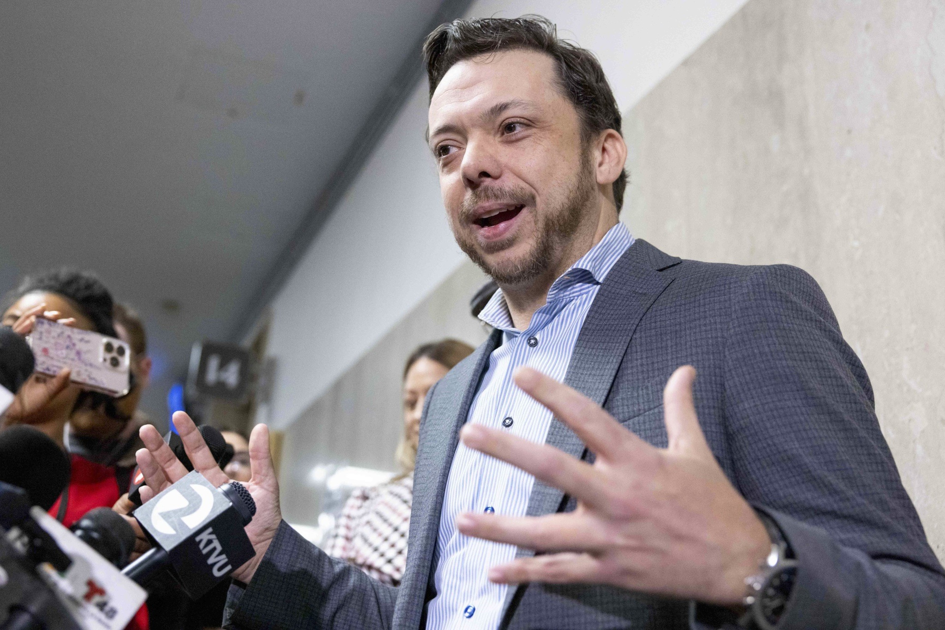 A man in a suit speaks animatedly to the press, gesturing with his hands. Multiple microphones are held up to him, and others stand around photographing.