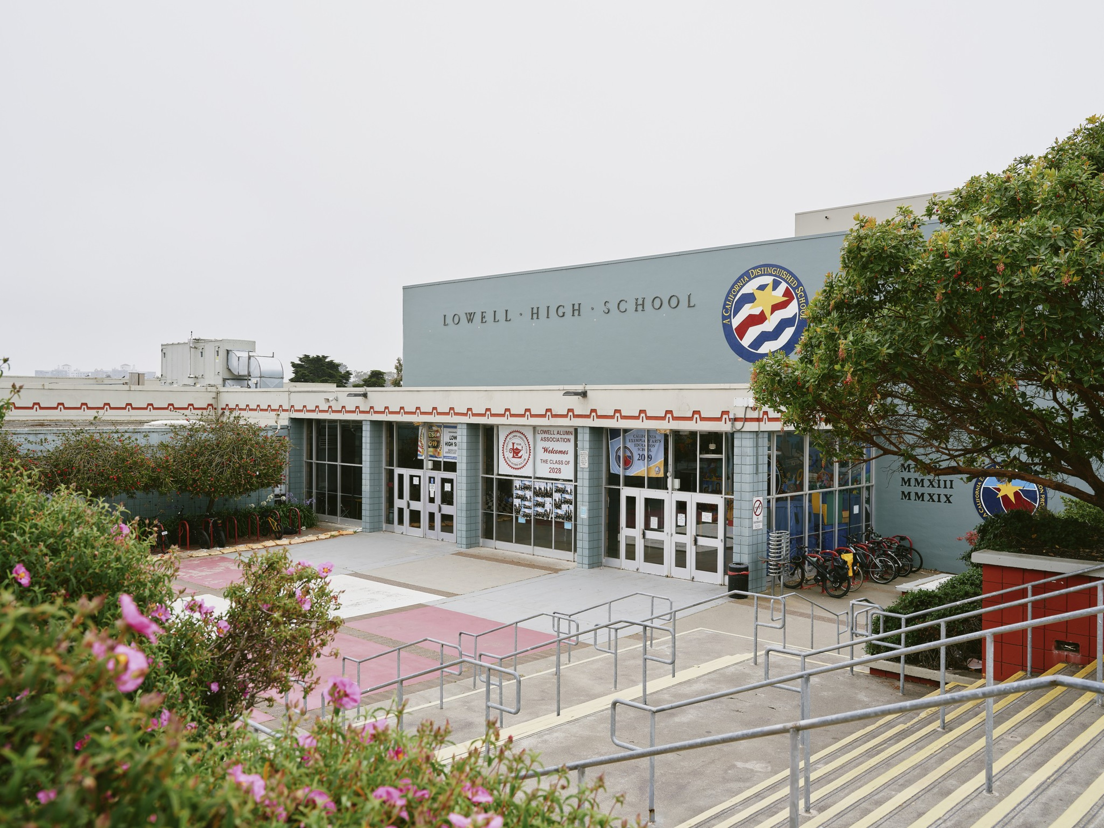 The image shows the entrance of a high school with glass doors, a large school emblem, and signs. There are bikes parked on the right, with trees and flowers nearby.