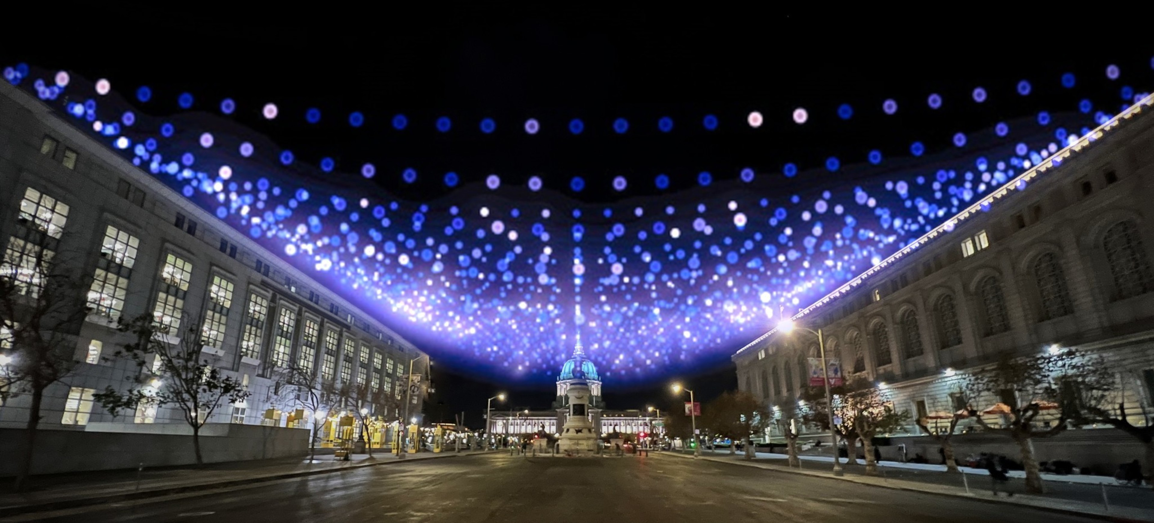 A nighttime street scene features two large buildings and a dome lit blue in the center. Overhead, a canopy of blue and white lights creates a wavy pattern.