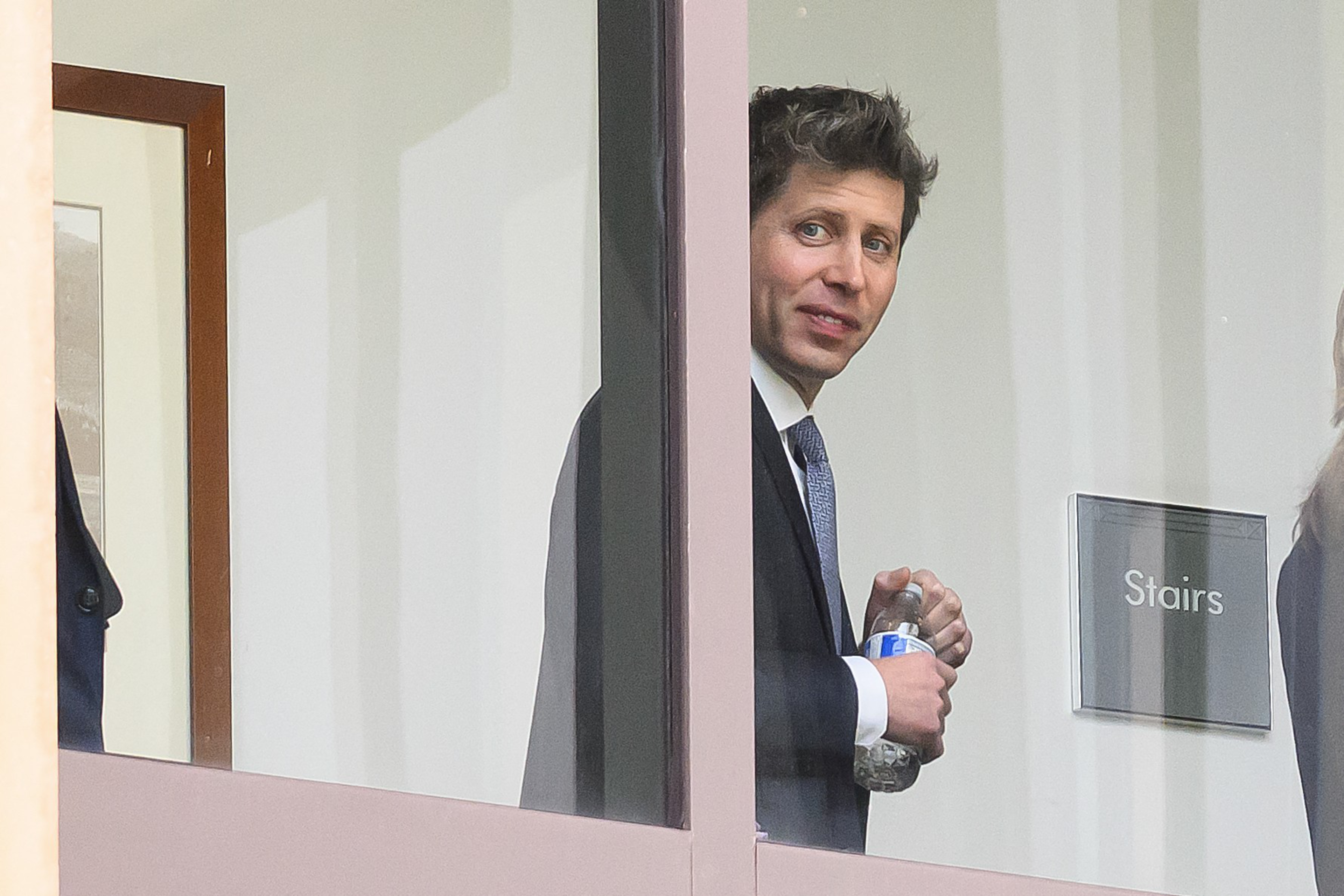 A man in a suit holding a water bottle stands inside a building near a door labeled “Stairs,” looking through a glass panel.