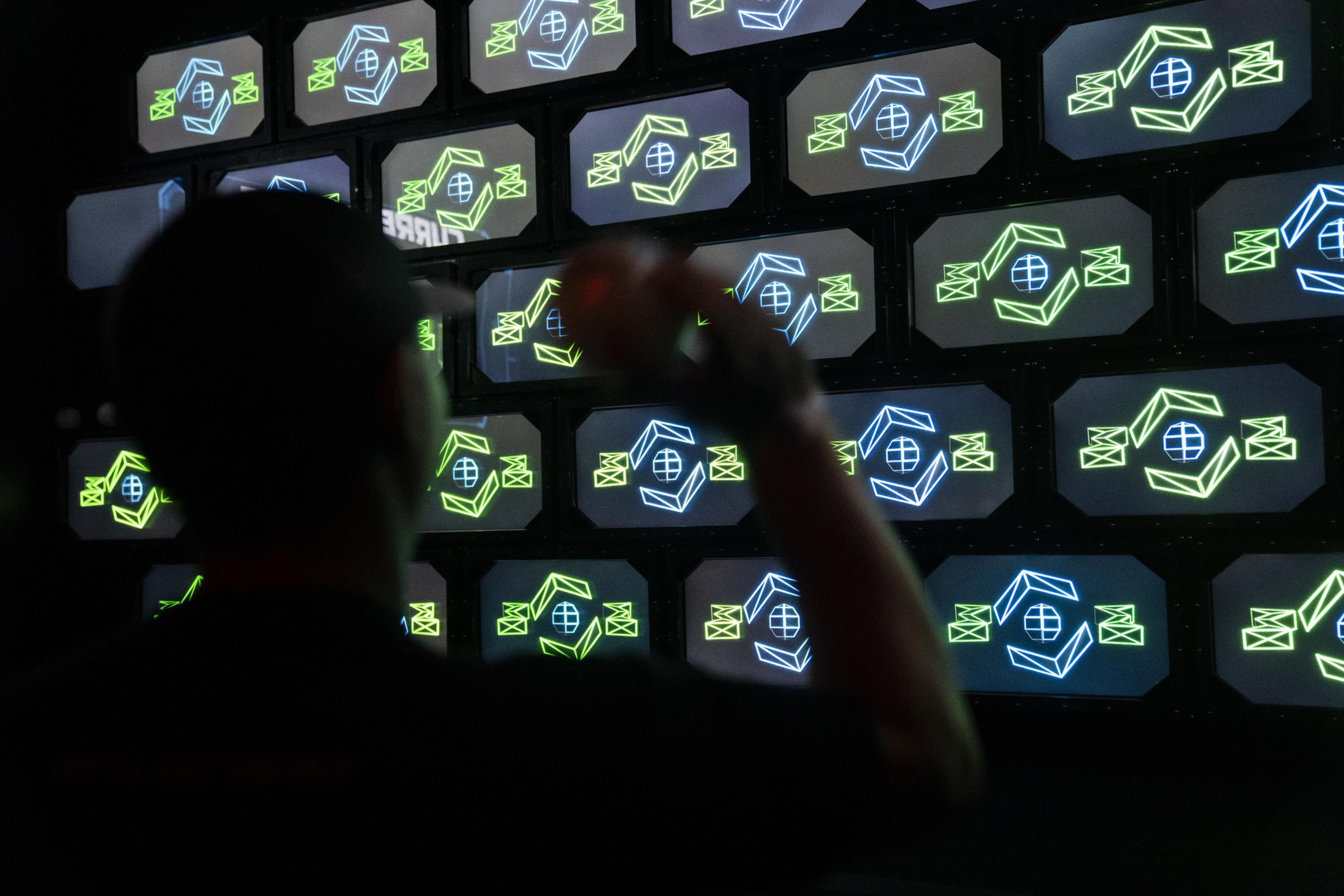 A silhouetted figure stands in front of a wall of futuristic screens displaying neon geometric symbols in blue and green.