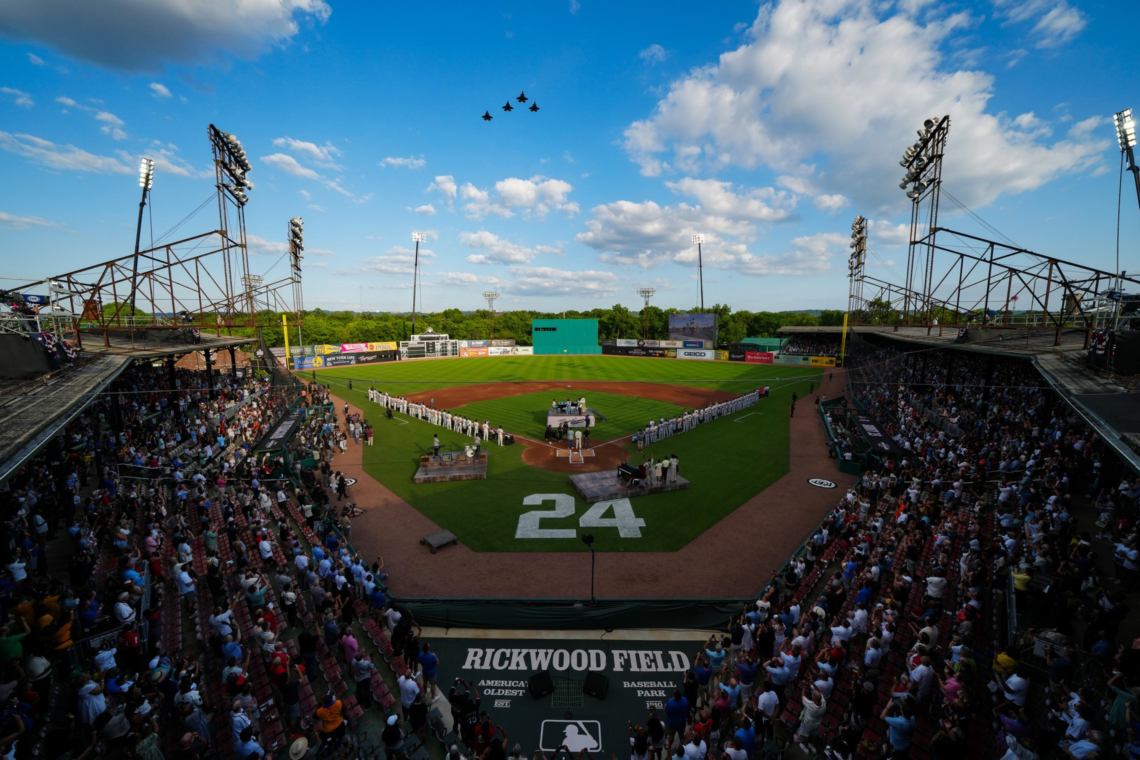 The image shows a baseball field with teams lined up on either side of home plate. A large number 24 is displayed on the field, and jets are flying overhead.