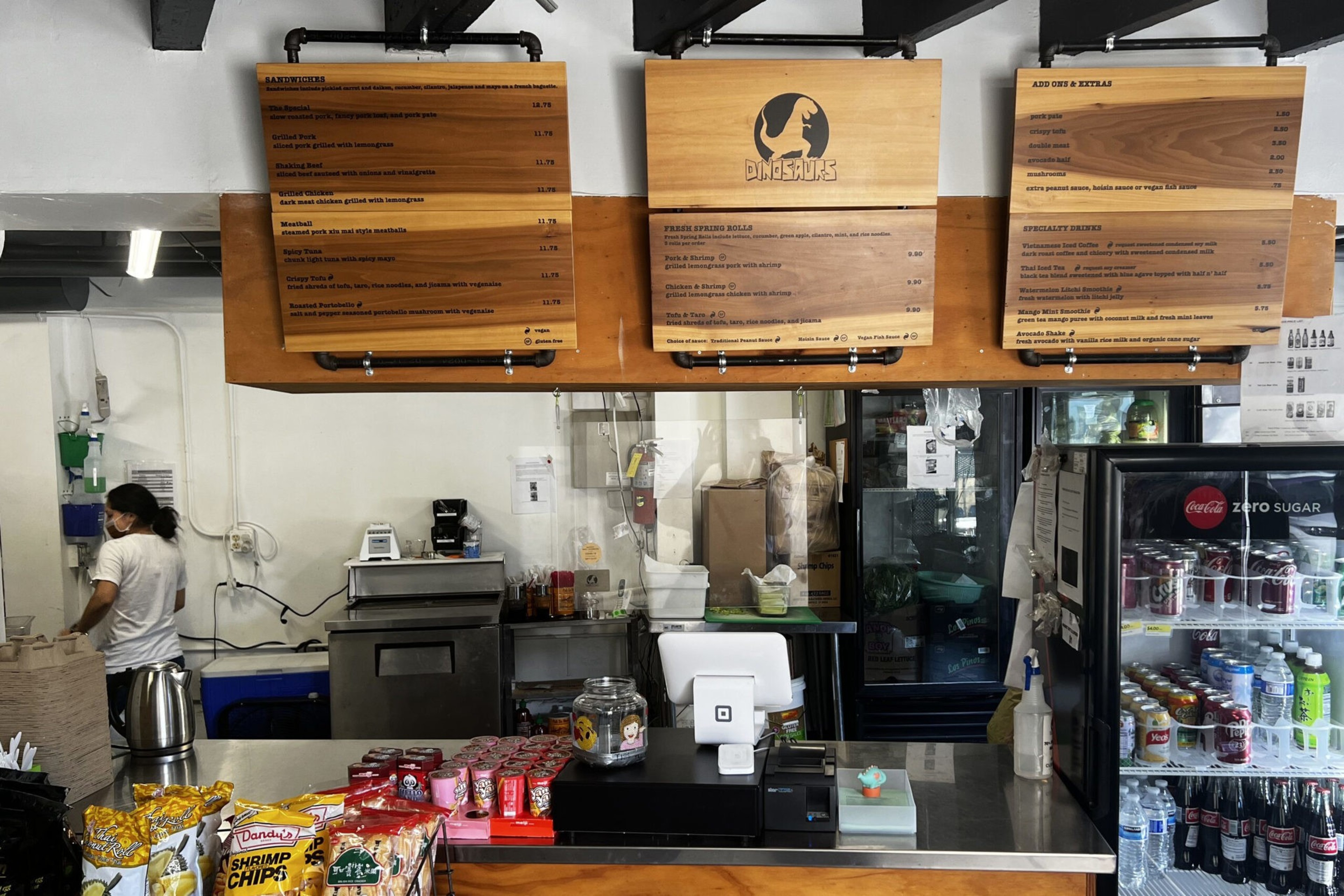 A restaurant counter with wood menu boards above, a cashier register, snacks like shrimp chips and drinks in a refrigerator, and a worker behind the counter.