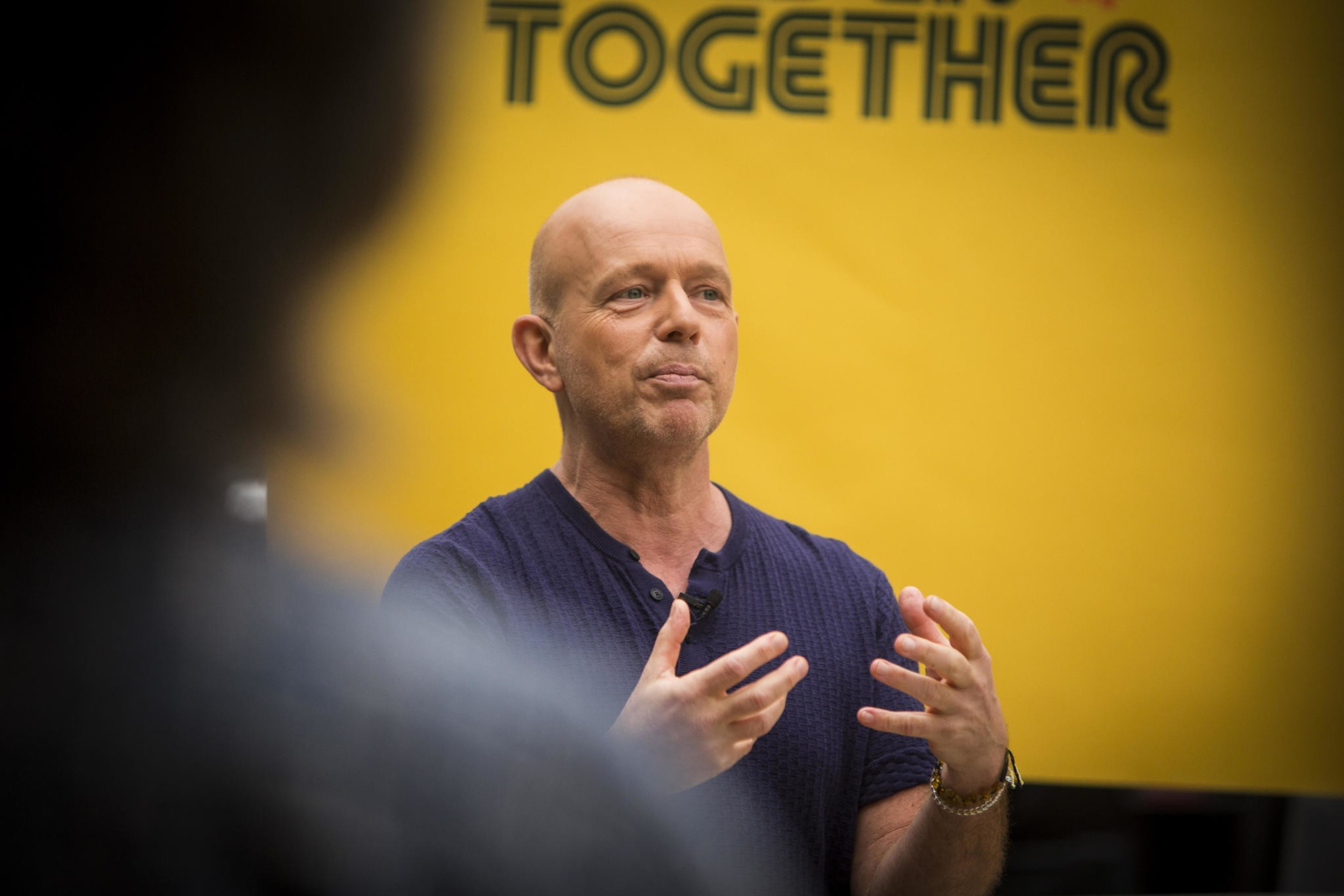 A bald man in a dark shirt gestures with his hands while speaking in front of a yellow background with partially visible text reading &quot;TOGETHER.&quot;