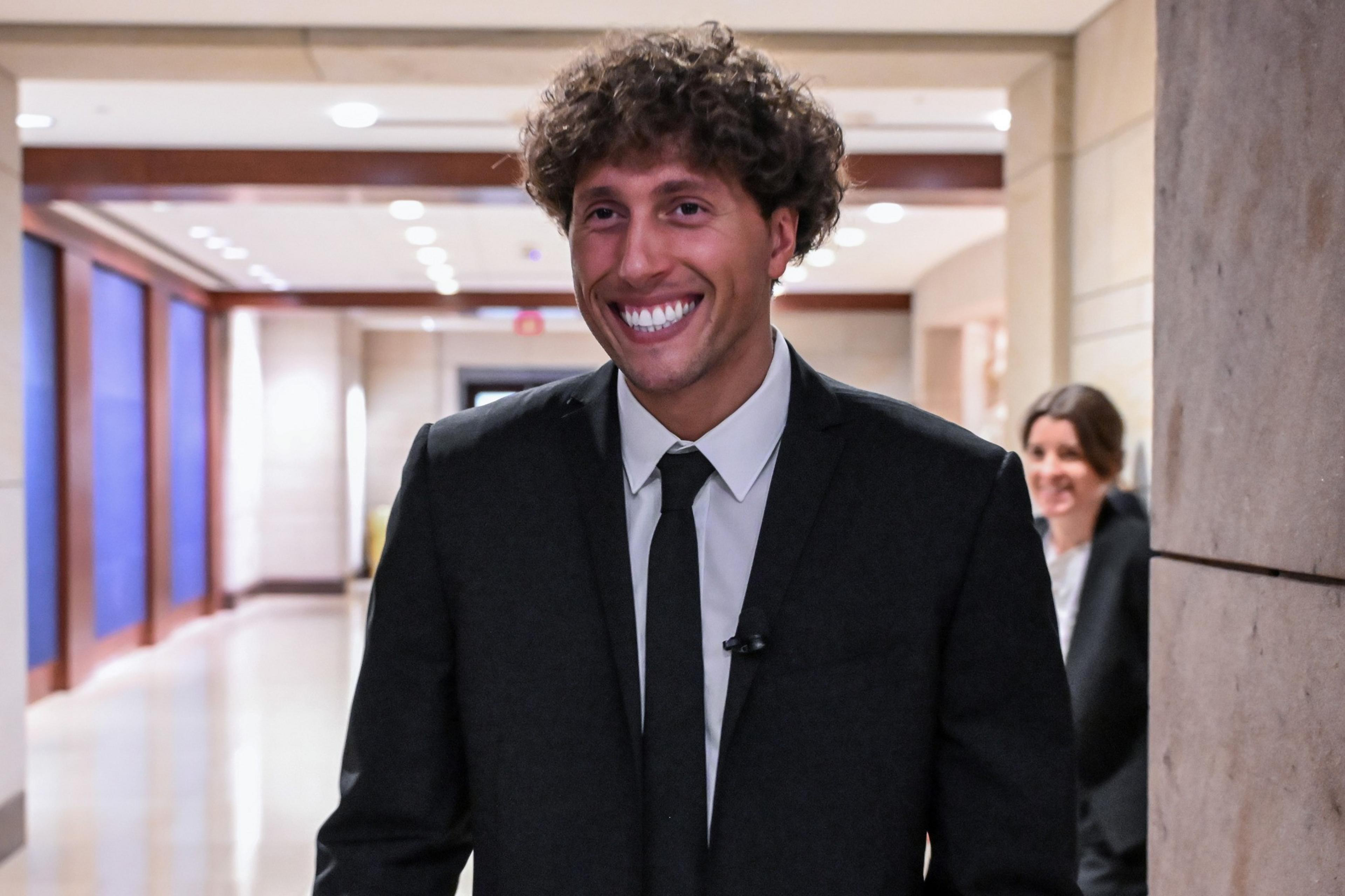 A person with curly hair is smiling widely in a hallway, wearing a dark suit and tie. The background shows lights and a person in focus behind him.