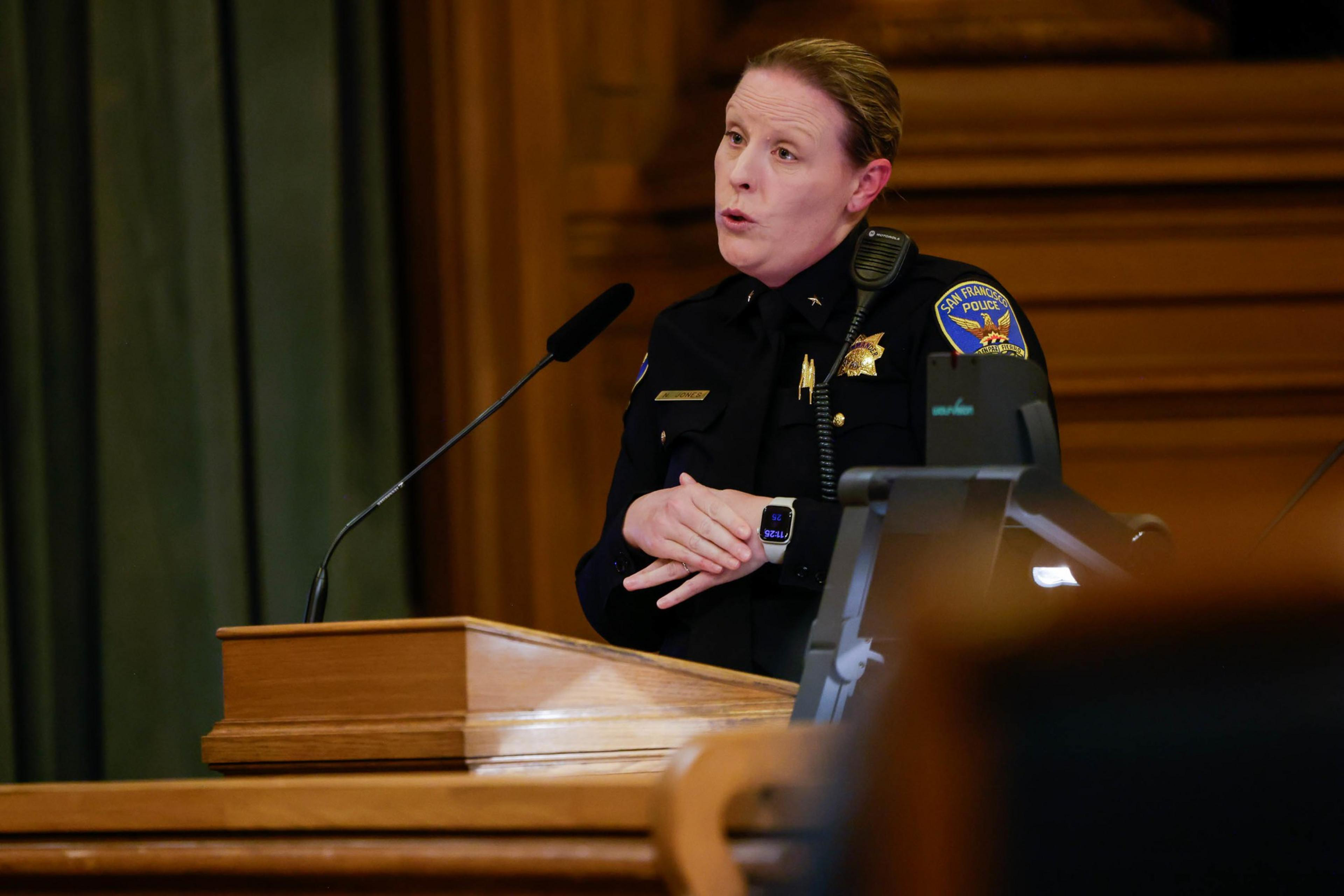 A female police officer in uniform speaks at a wooden podium, with a microphone in front and a police badge on her shoulder.