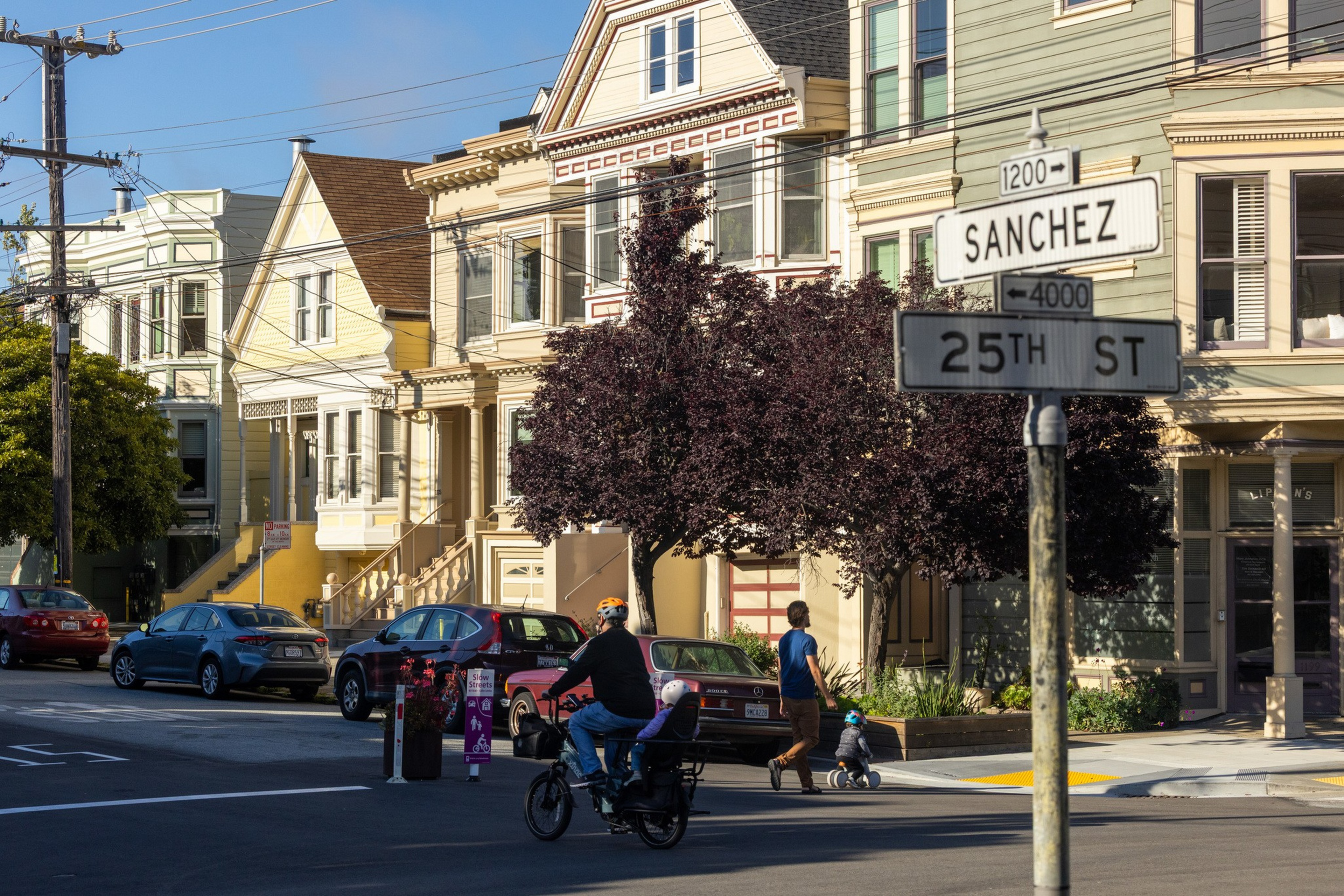 A cyclist with a helmet rides a bike with a child seat past a pedestrian and toddler on a scooter near a street sign reading Sanchez and 25th St.