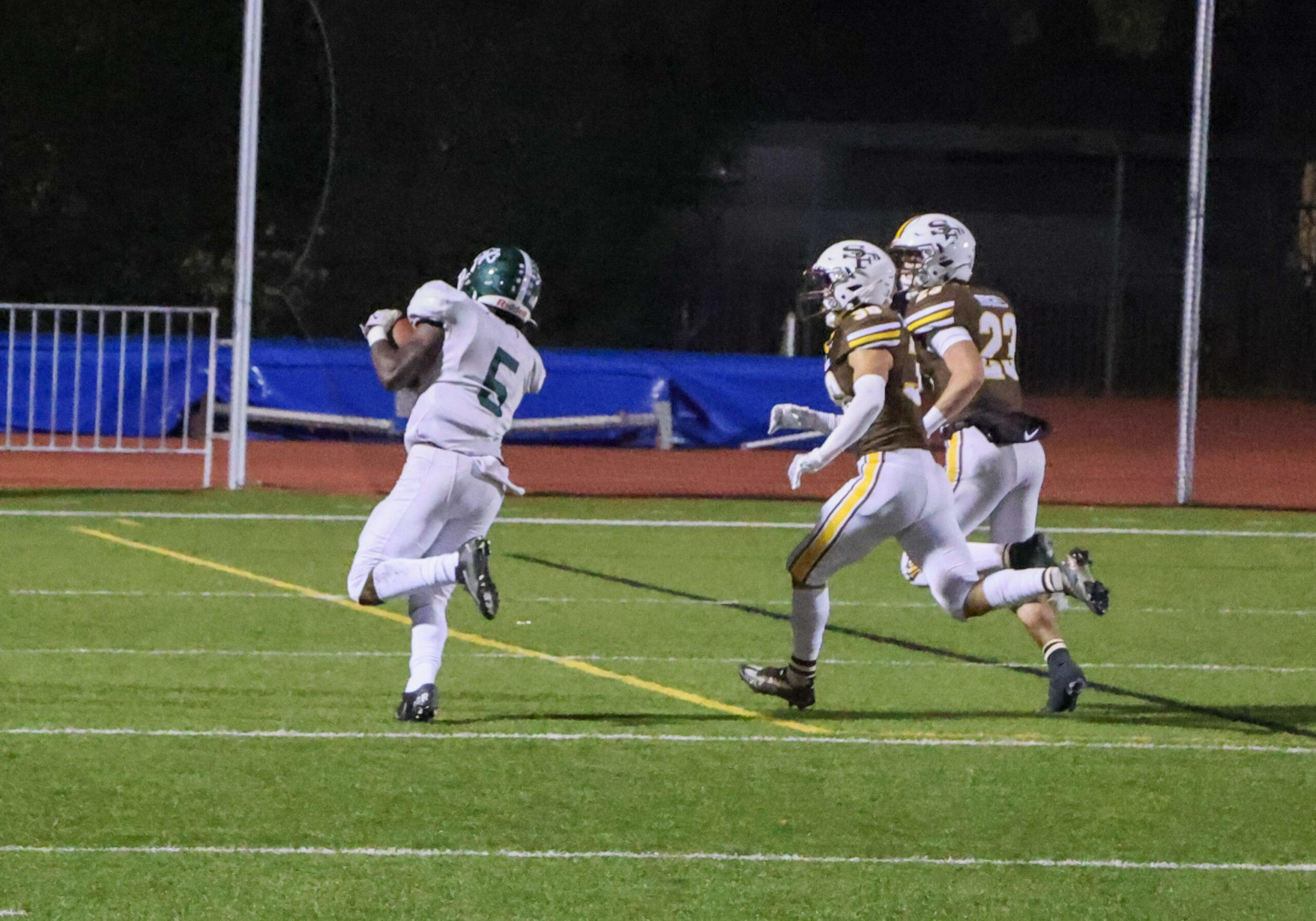 A football player in a white uniform catches the ball while two opposing players in brown and white uniforms run closely behind him on a green field.