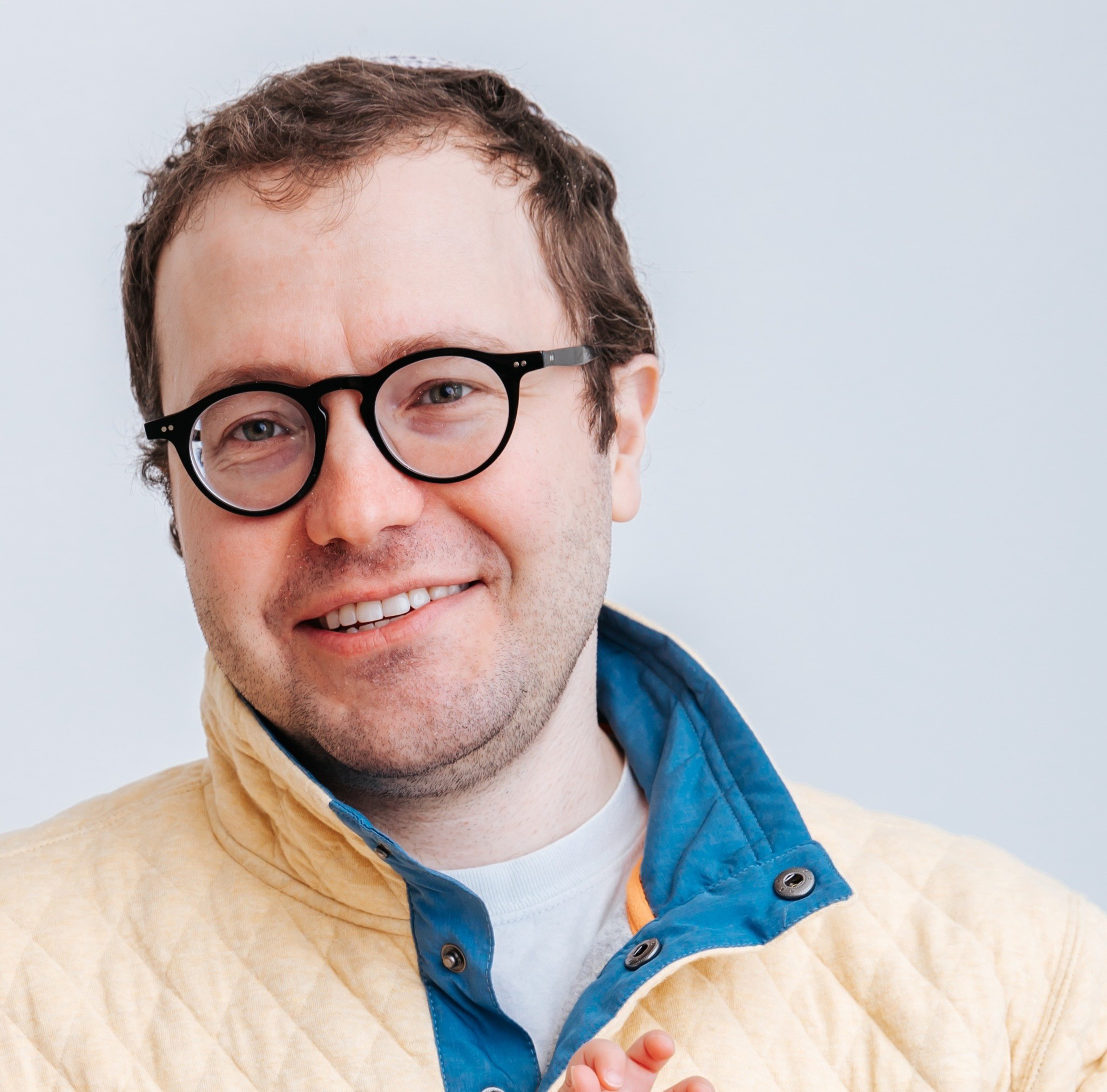 A smiling man with short brown hair and black round glasses wears a beige quilted jacket with a blue collar over a white shirt against a plain background.