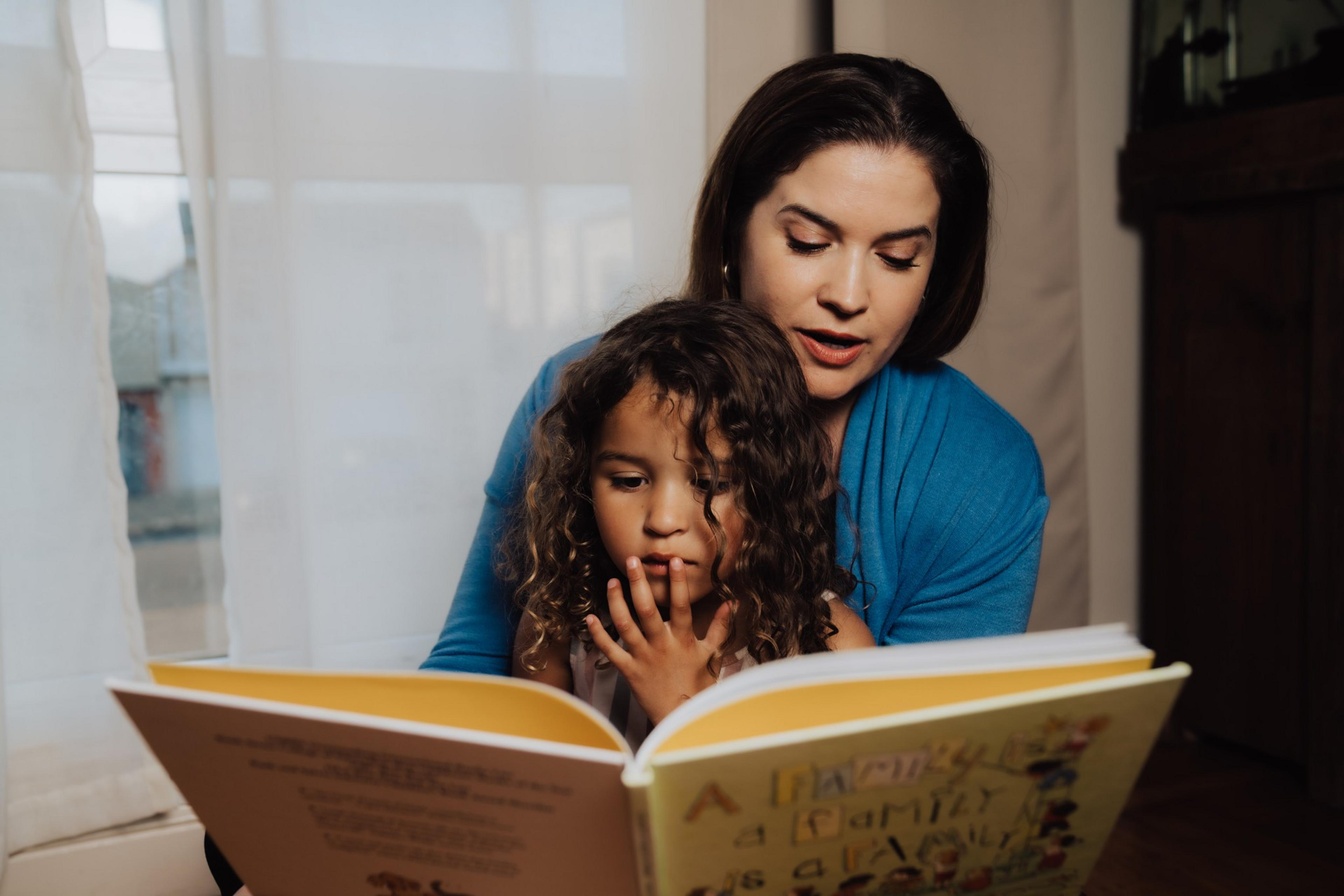 A woman in a blue sweater reads a colorful book to a young girl with curly hair. They sit closely together in a softly lit room.