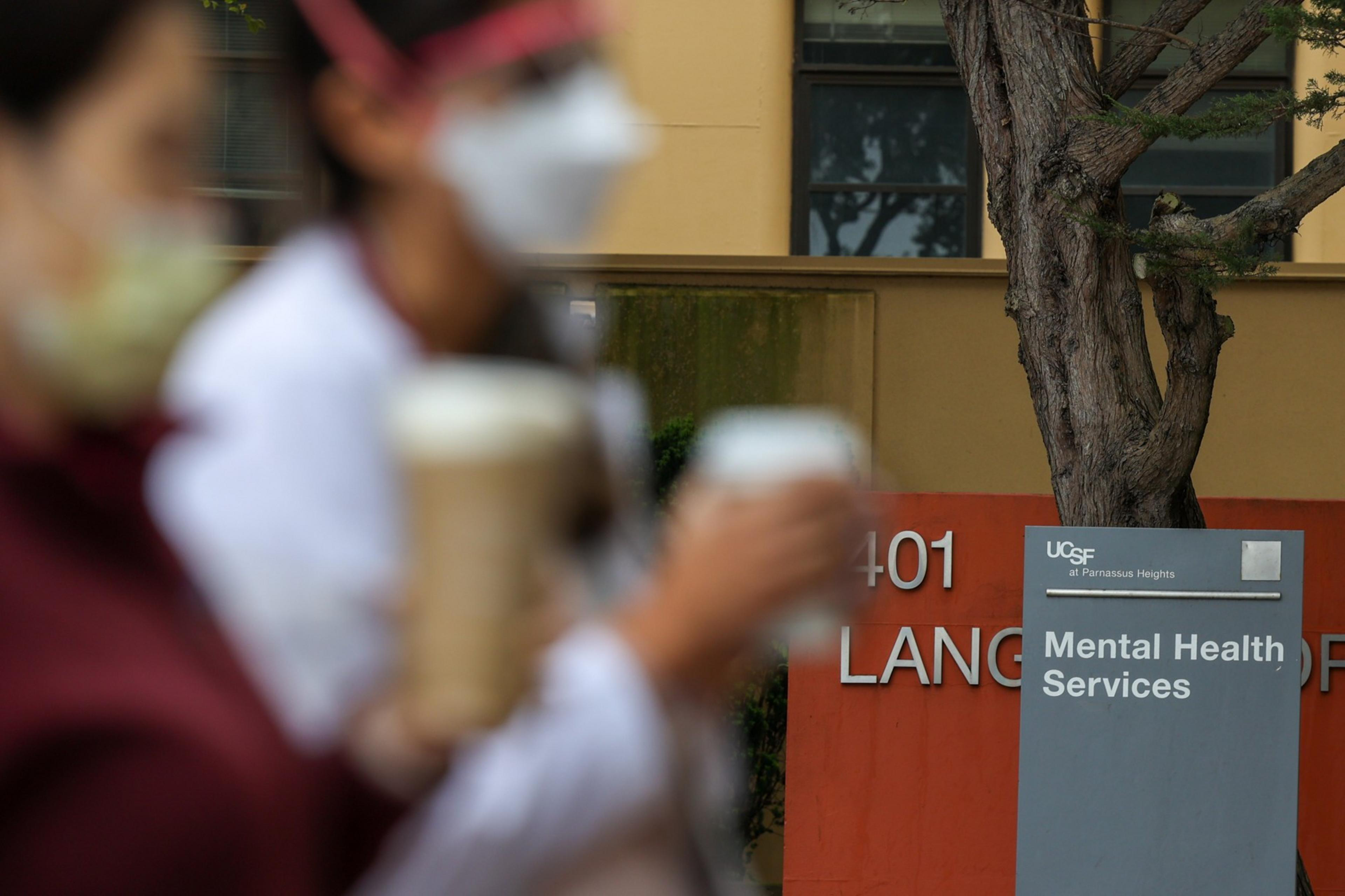 The image shows two blurred individuals in masks holding drinks in front of a "Mental Health Services" sign and a tree, with a building in the background.