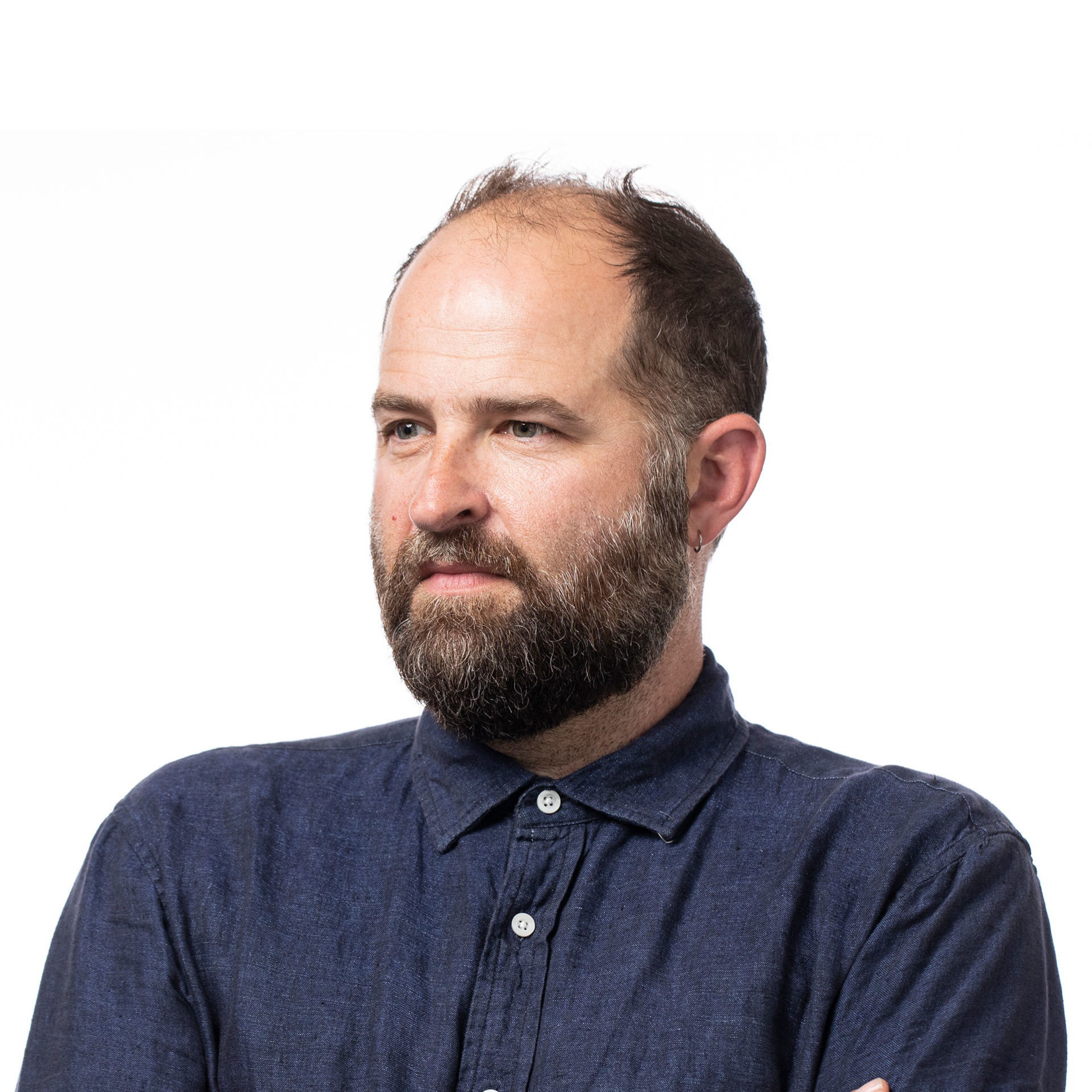 A bearded man with short, thinning brown hair wears a dark blue button-up shirt, looking thoughtful with arms crossed against a white background.