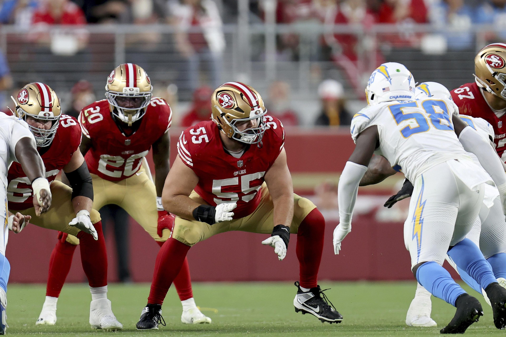 Football players from the San Francisco 49ers in red and gold jerseys face off against Los Angeles Chargers players in white and blue uniforms on the field.