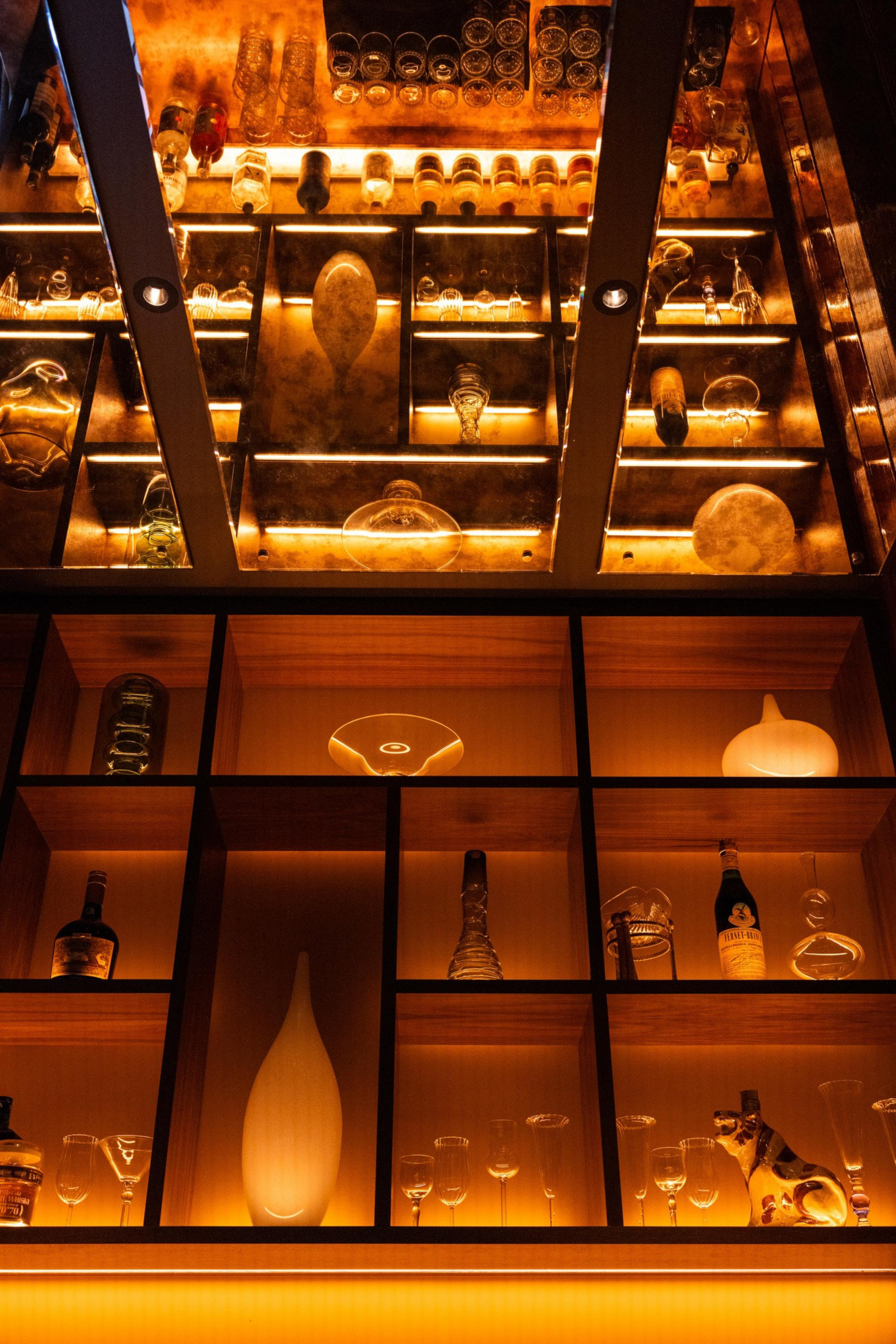 Glass shelves with warm backlighting display various glassware, bottles, a gourd-shaped vase, and a small ceramic dog figurine below more glass items above.