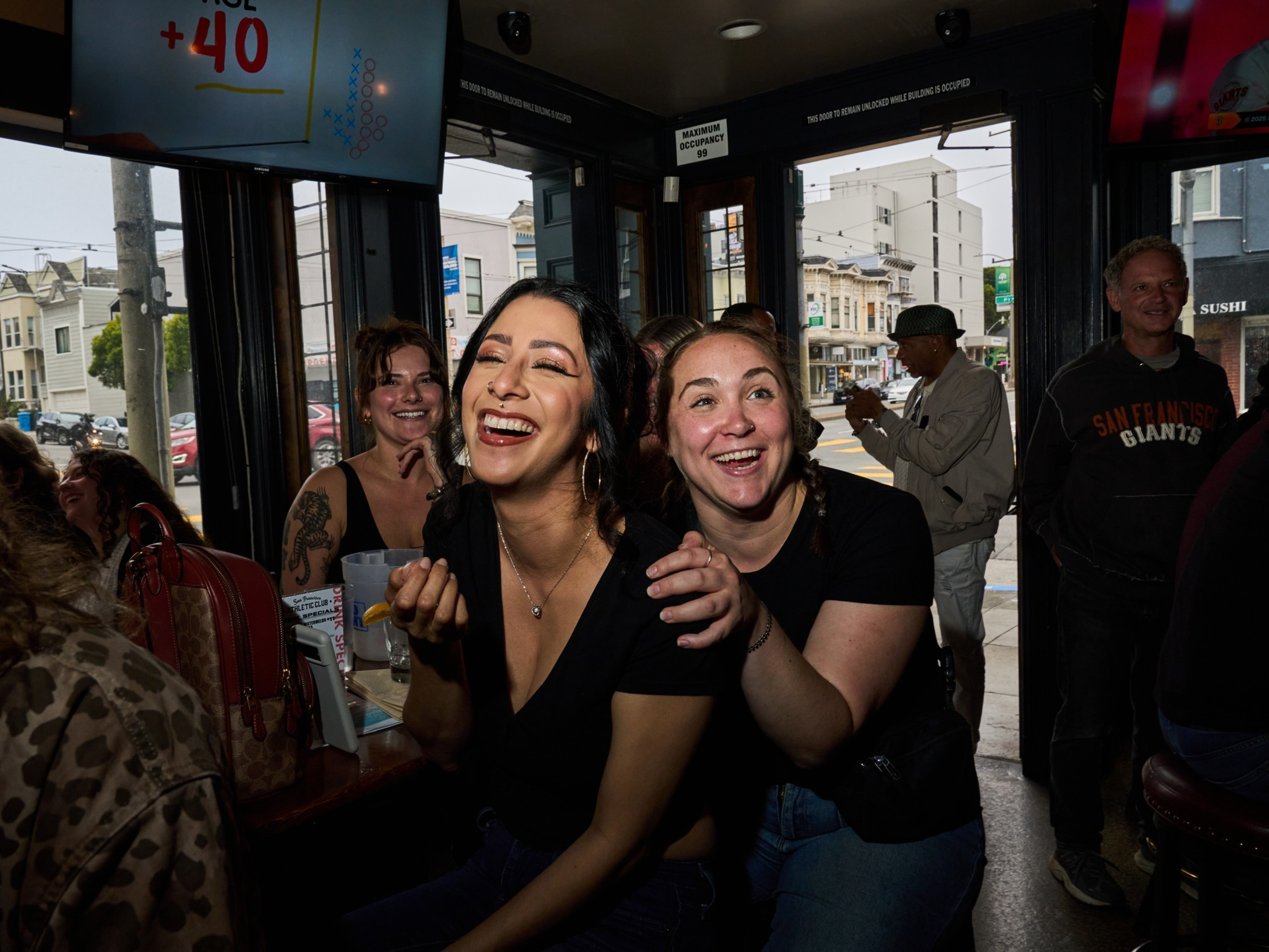A joyful group of people is gathered in a lively bar. Two women in the center are laughing heartily, while others around them also appear cheerful. The bar is bustling and cozy.