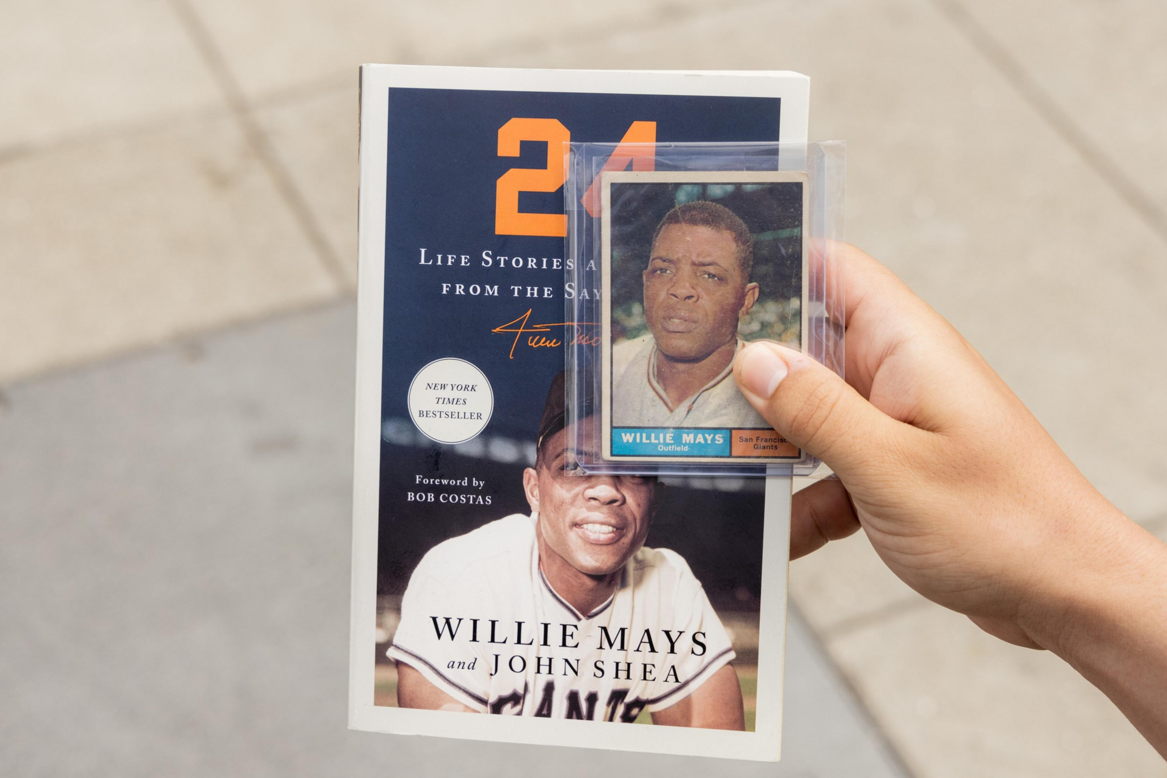 A hand is holding a book titled &quot;24: Life Stories and Lessons from the Say Hey Kid&quot; by Willie Mays and John Shea, along with a plastic-encased baseball card of Willie Mays.