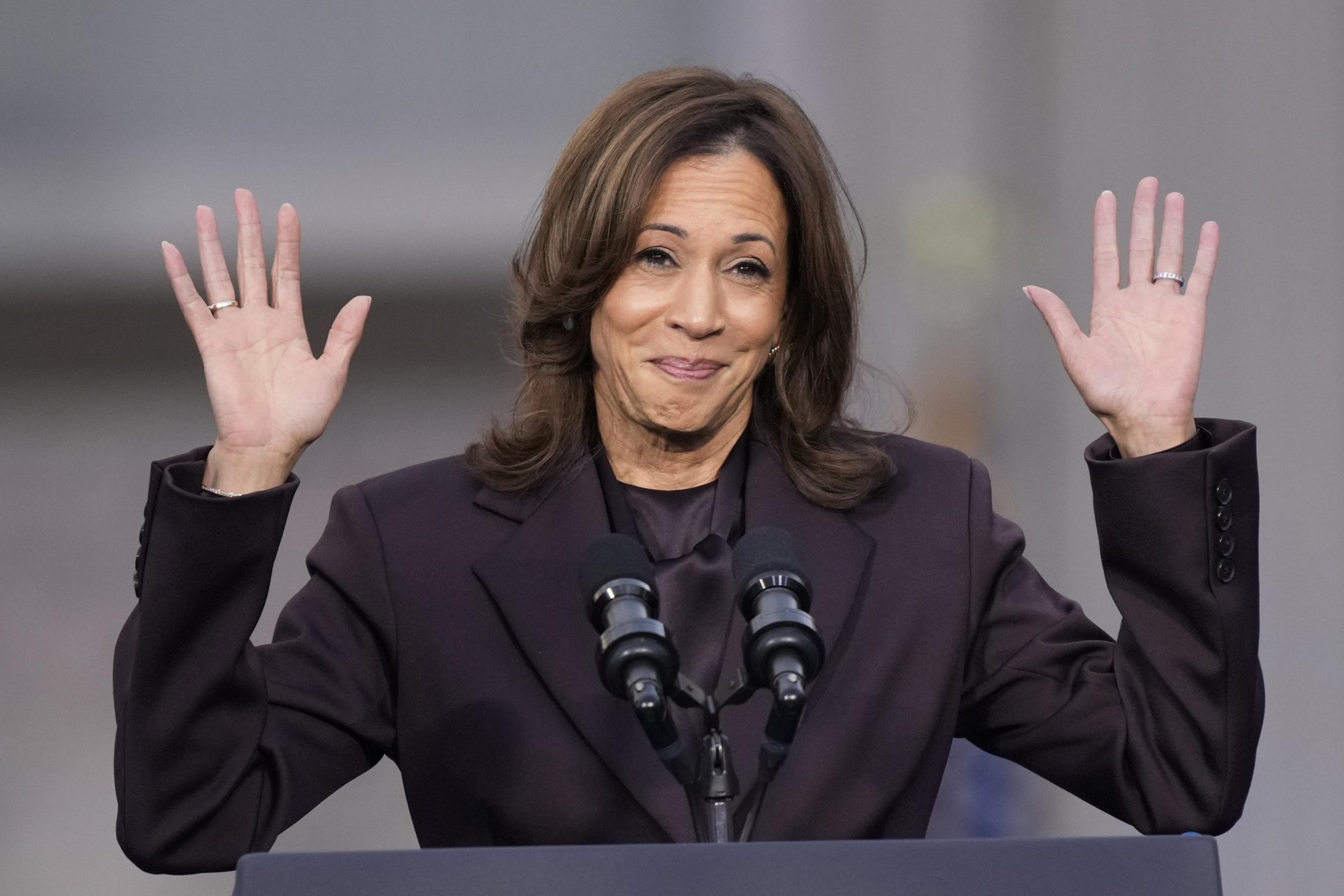 A woman stands at a podium with a microphone, wearing a dark suit, smiling, and raising both hands.