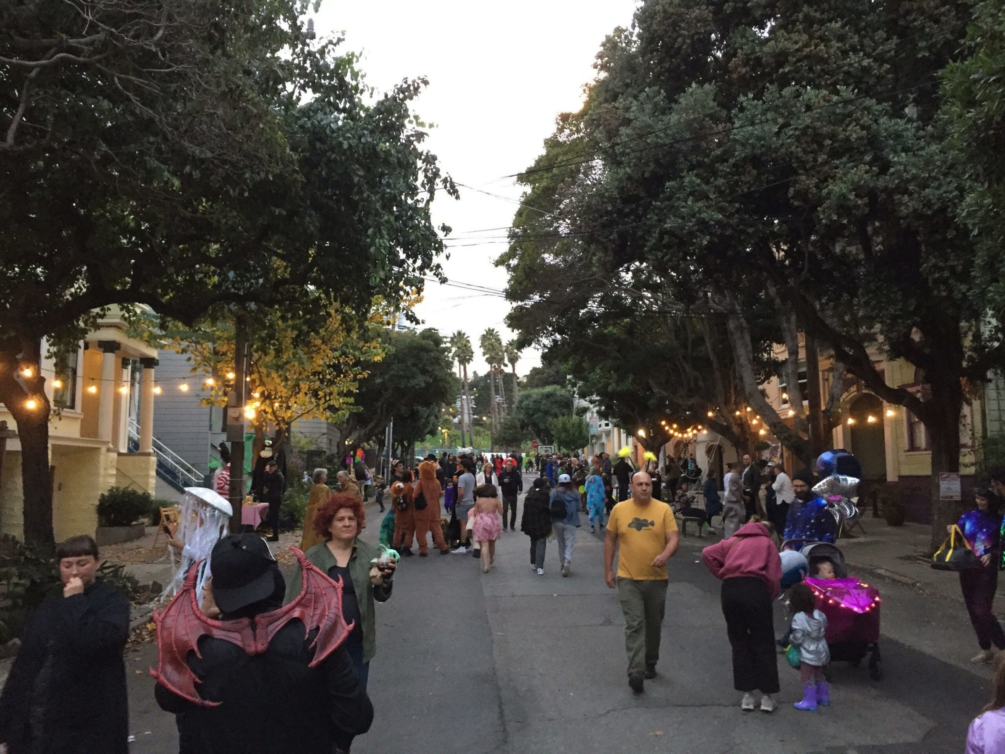 People of all ages dressed in various costumes walk down a tree-lined street decorated with string lights during dusk.