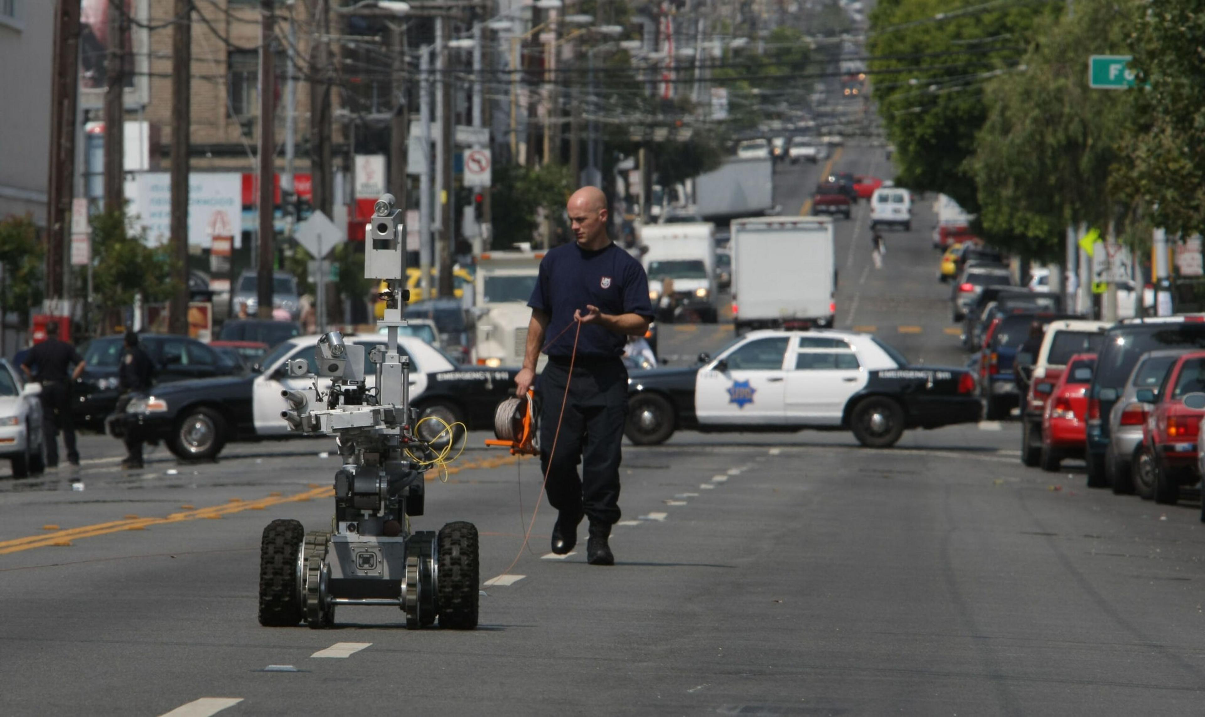 A man in a dark uniform walks behind a bomb disposal robot on a city street blocked by police cars.