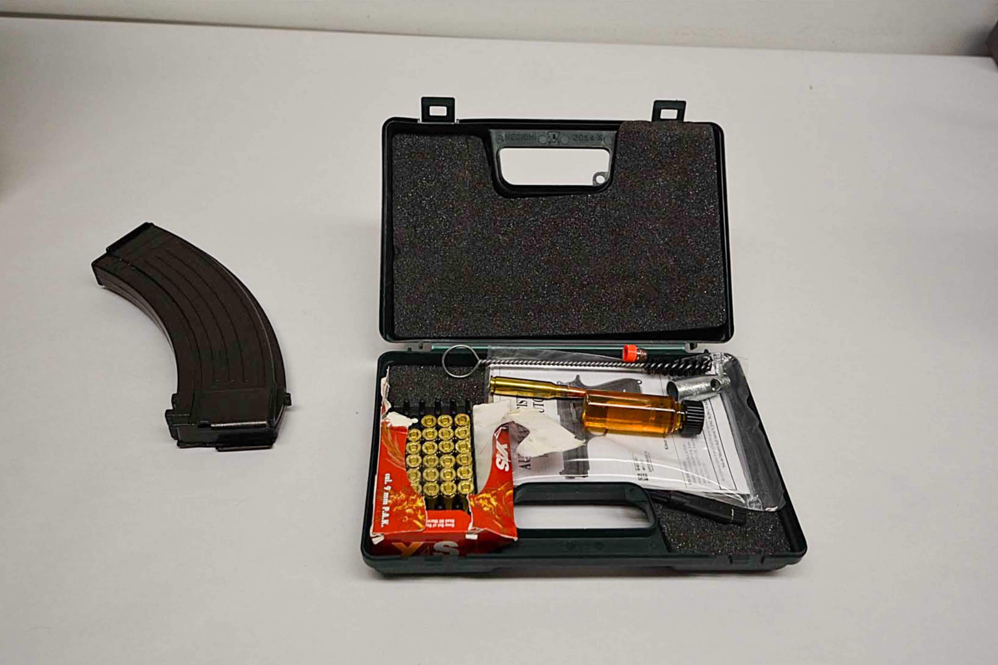 A pistol cleaning kit with brushes, oil, and paper, along with a detachable curved rifle magazine, are arranged on a white surface.
