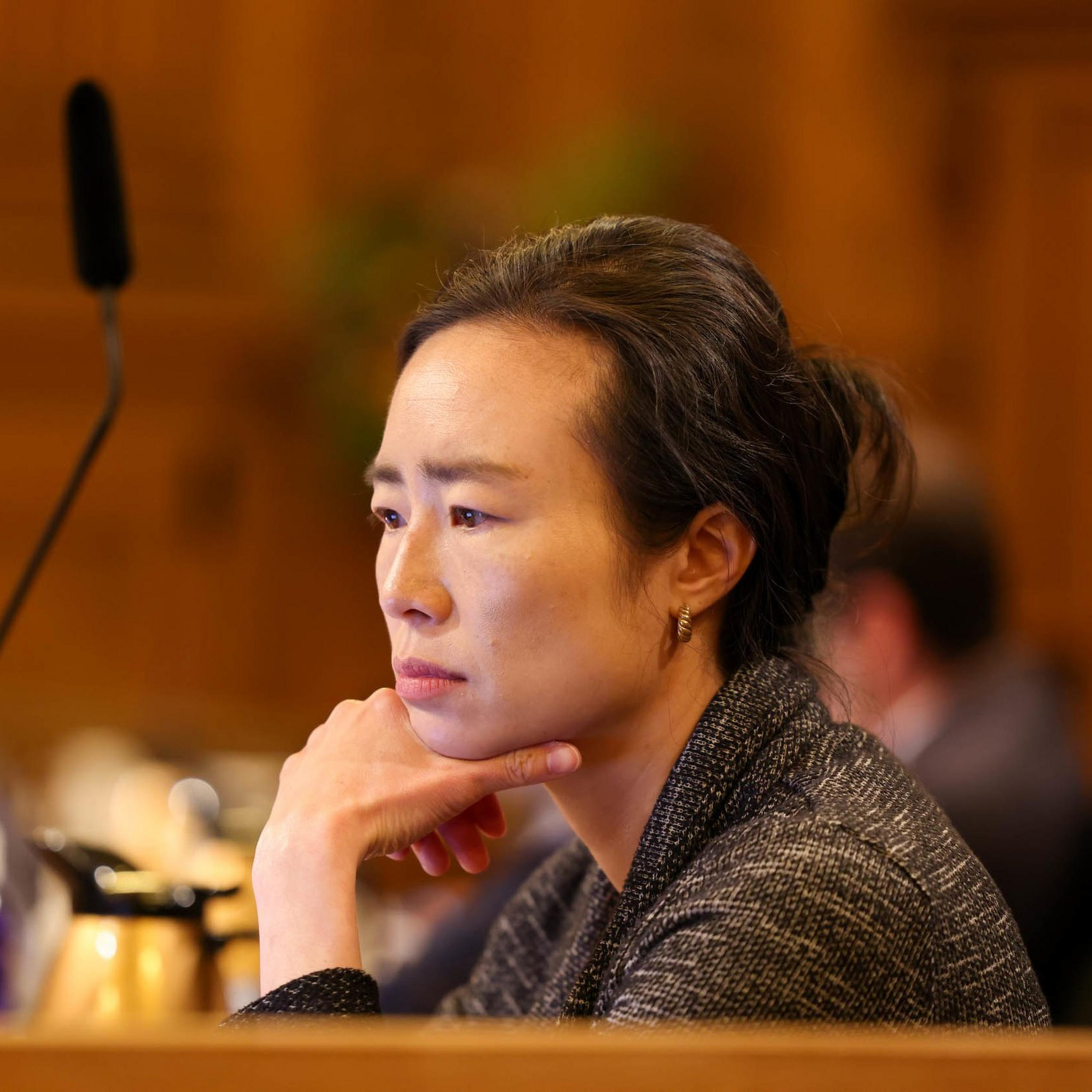 A person sits pensively with their chin resting on their hand. They appear engaged in thought. In the background are blurred figures and indoor lighting.