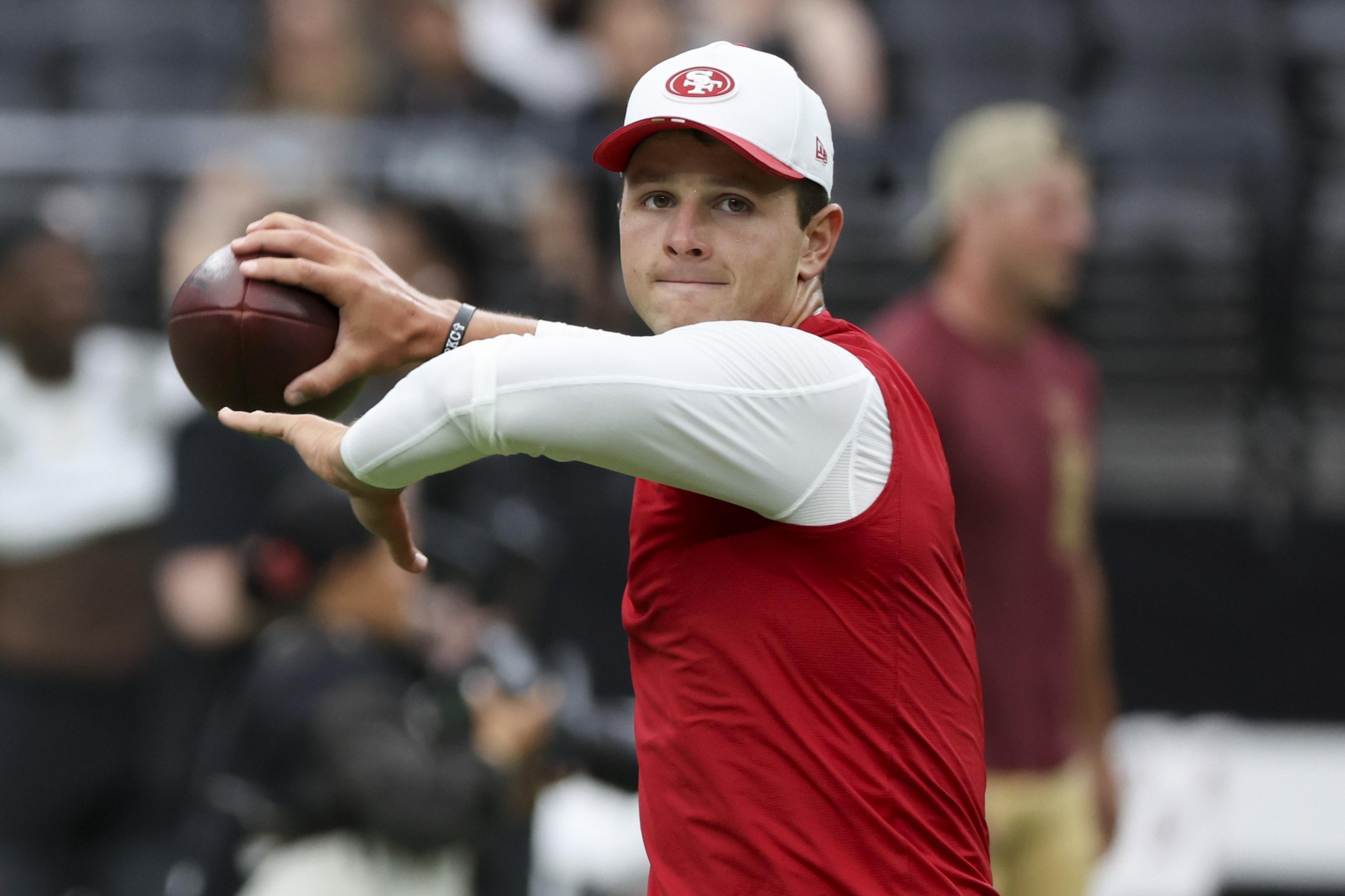 A man wearing a red and white San Francisco 49ers cap and red shirt is poised to throw a football during practice.