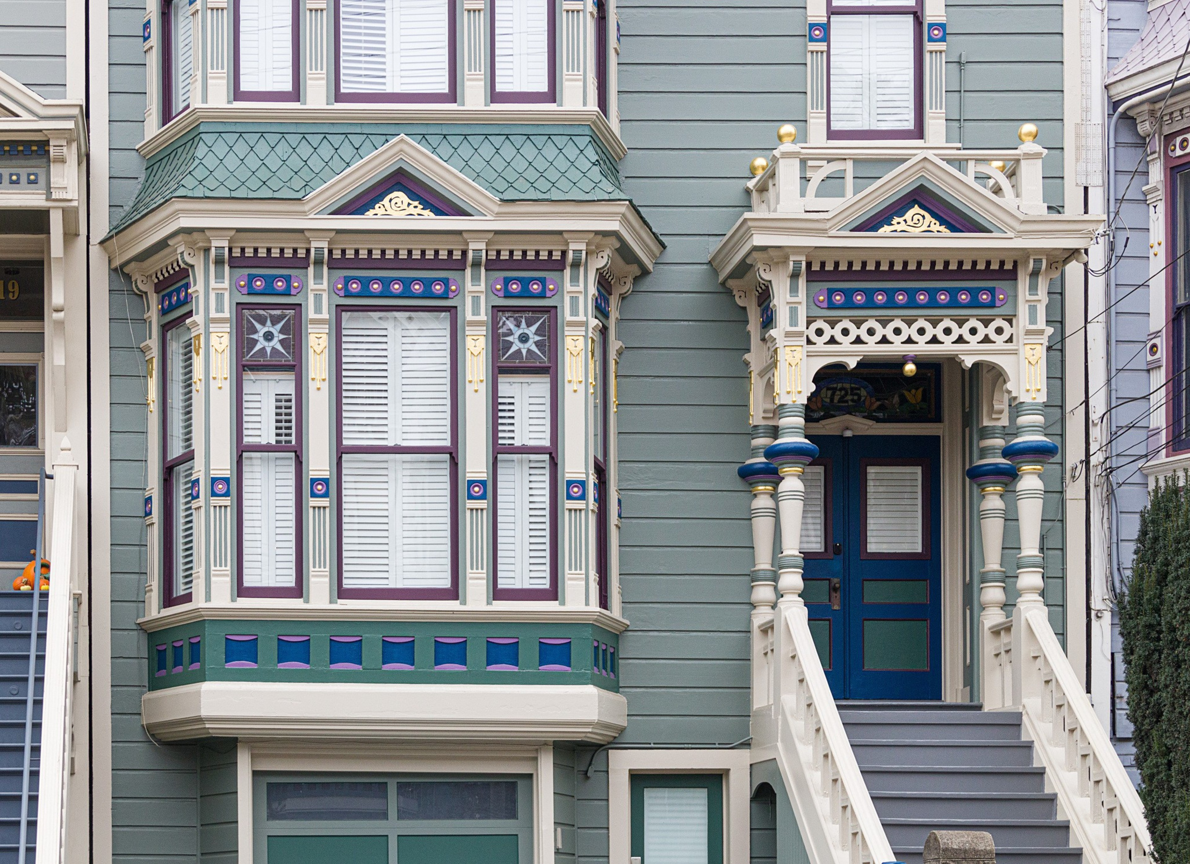 A detailed Victorian-style house facade in blue and white tones features ornate window shutters, decorative trim, and a porch with columns and stairs leading to a blue door.