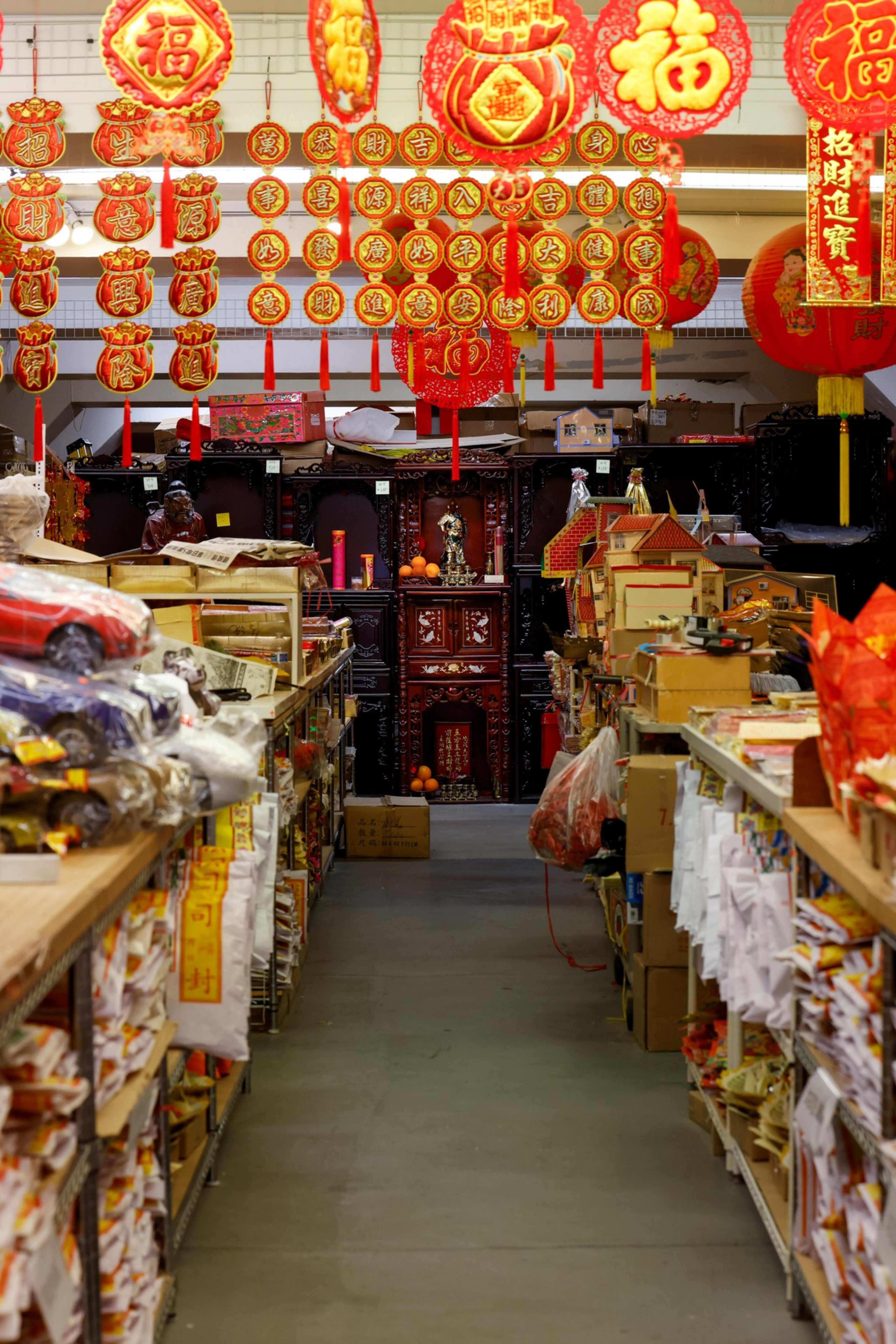 The image shows a store aisle filled with various items on shelves. Red and gold Chinese decorations and ornaments hang overhead, with a shrine visible in the back.