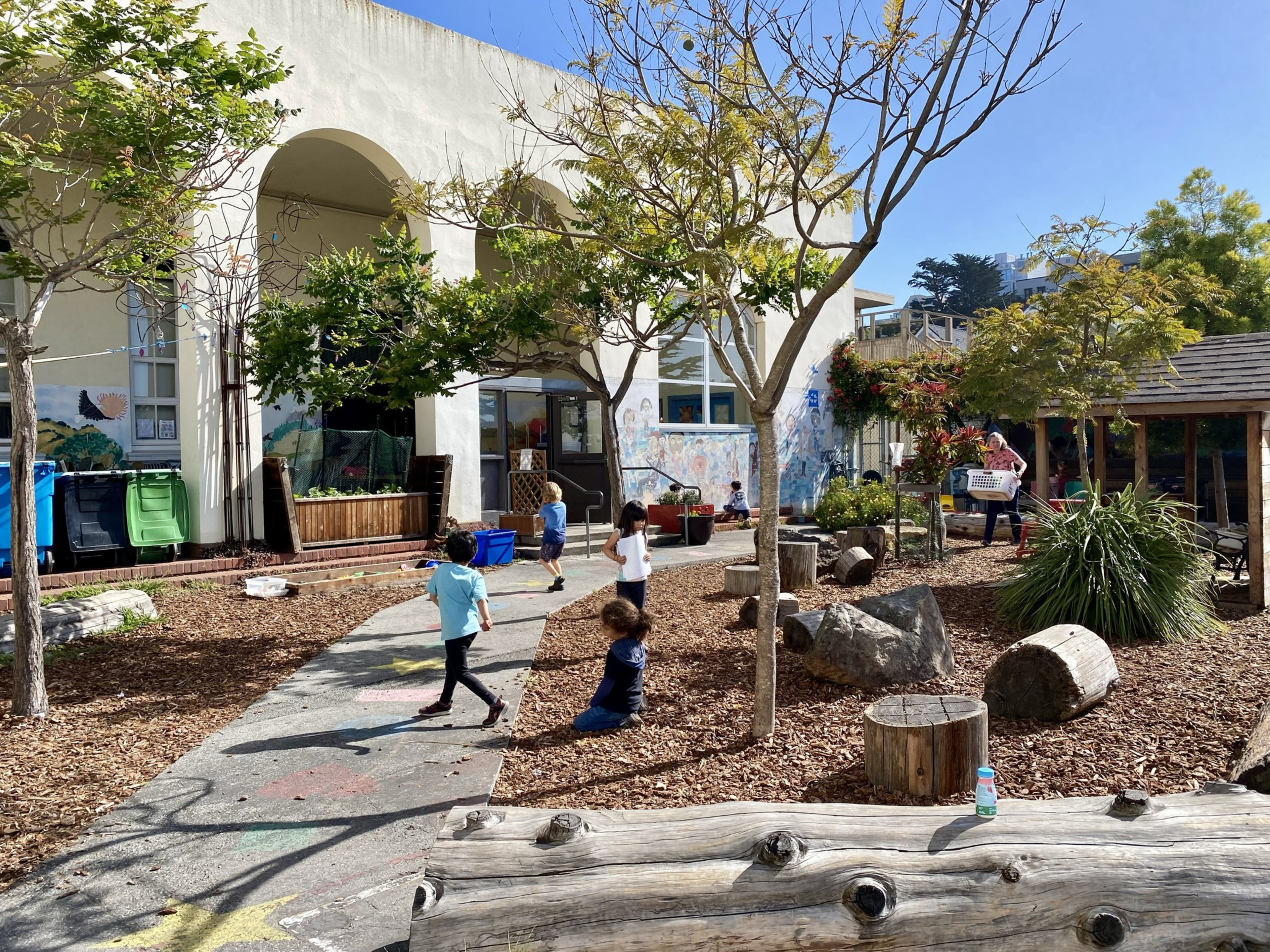 The image shows a sunny outdoor playground with children playing on a path amidst trees and logs, next to a building with colorful murals and recycling bins.