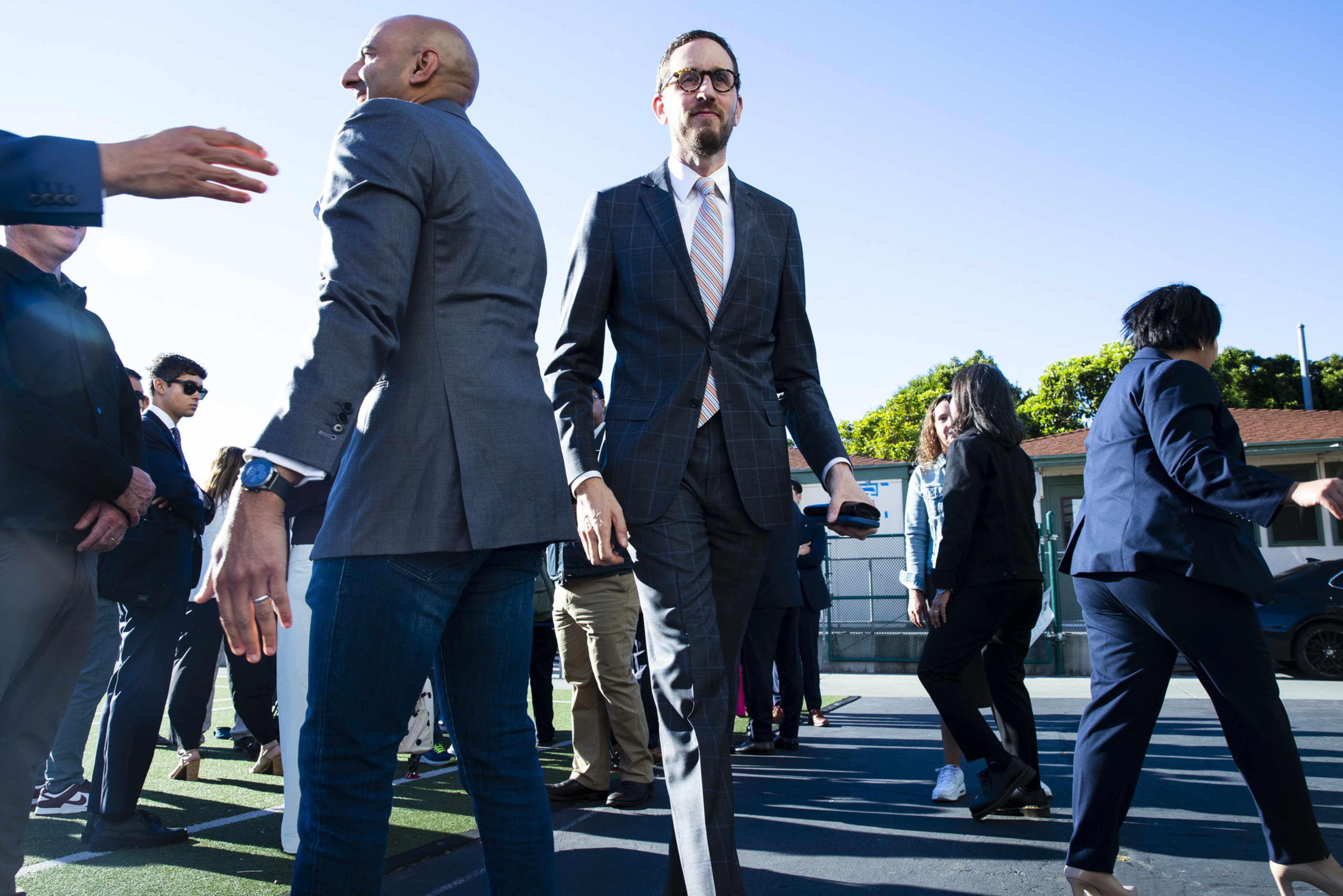 A group of professionally dressed people stand and walk outdoors under a bright blue sky, engaging in conversation or moving around an open area.