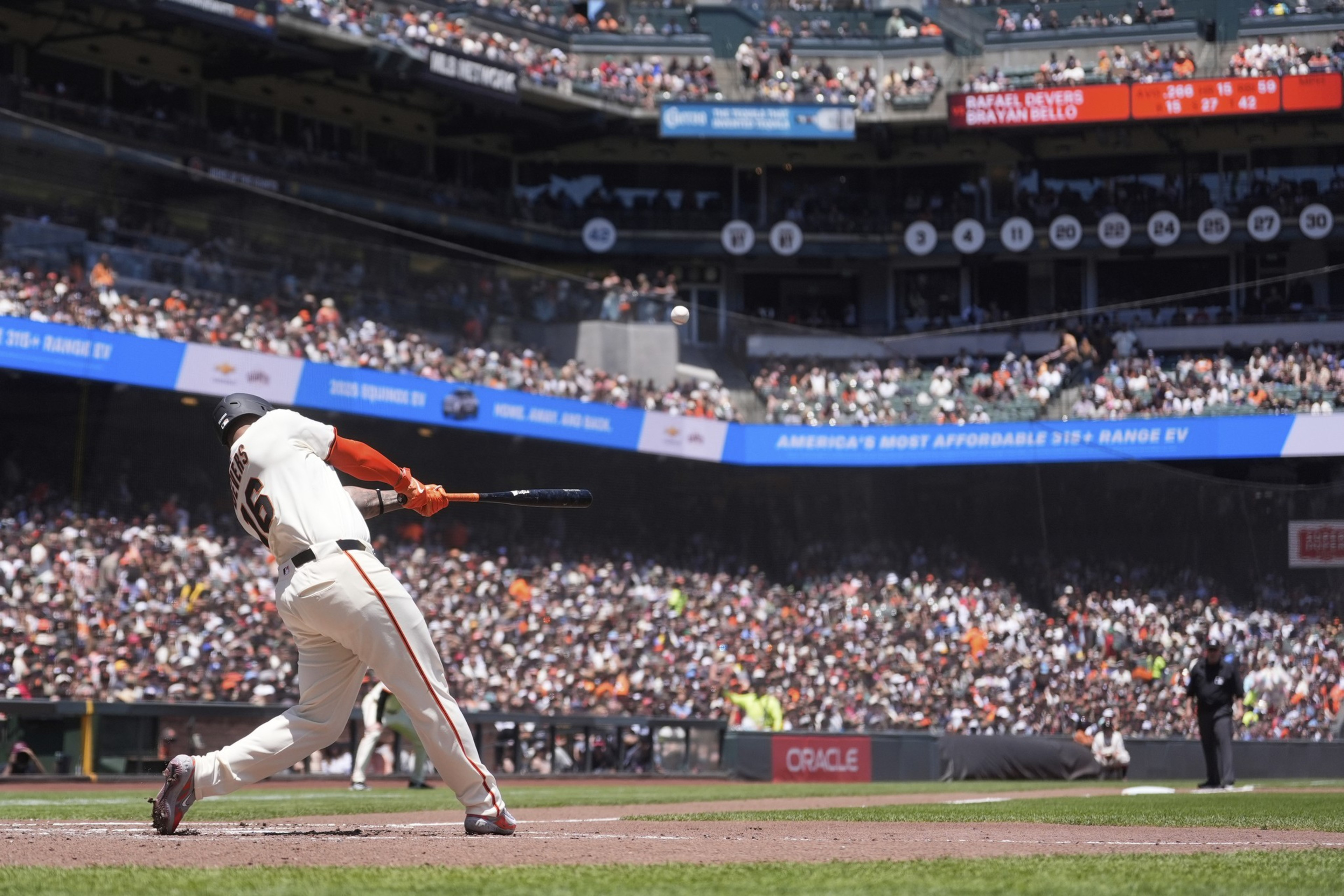 A baseball player in a white uniform with number 66 is mid-swing in a packed stadium. The crowd and scoreboards are visible in the background.