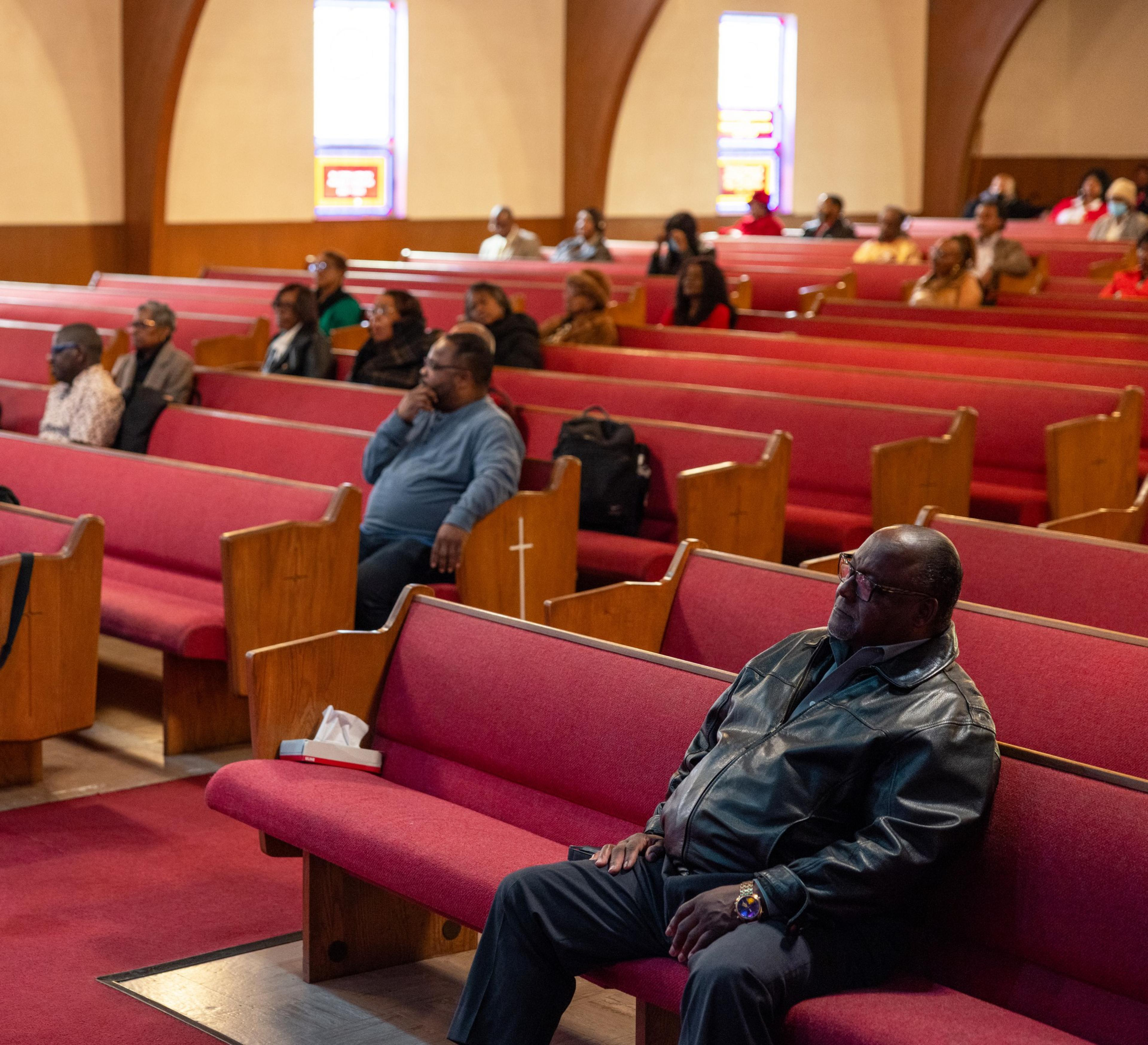 People are seated on red pews in a church, with wooden archways and stained glass windows in the background. The group appears to be listening attentively.