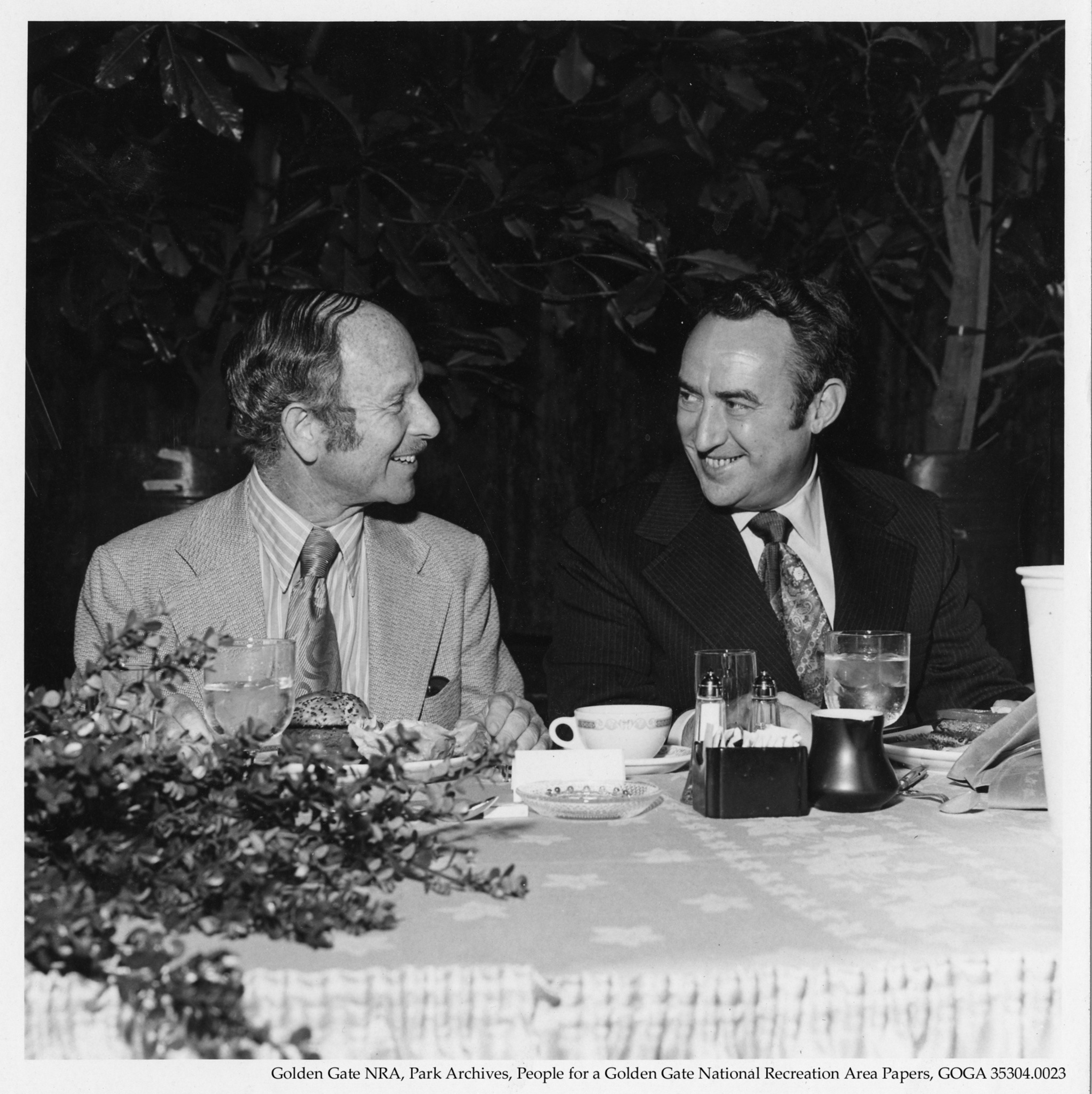 Two men in suits sit at a table outdoors, smiling at each other, with food, drinks, and tableware between them.
