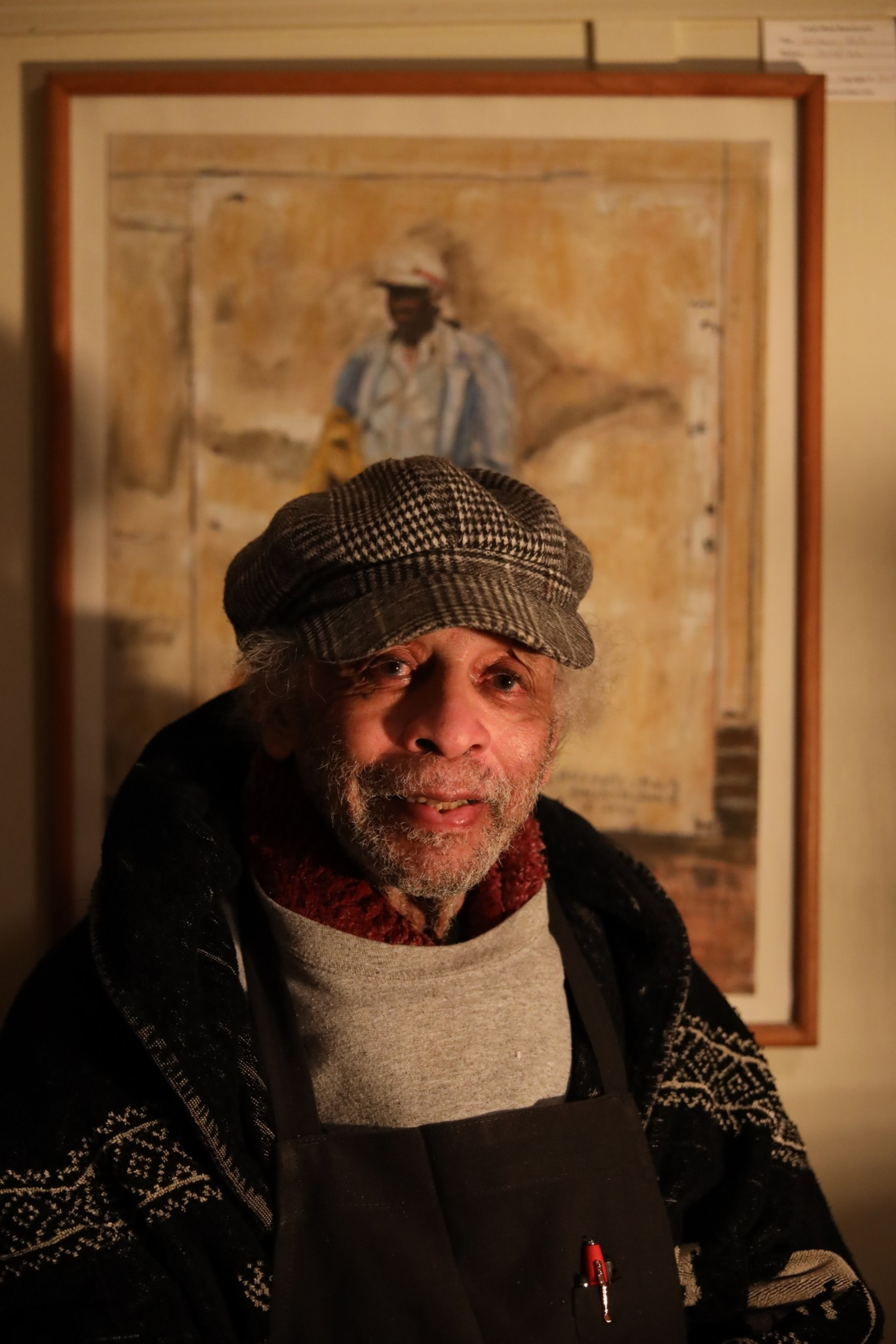 An older man with a graying beard wears a checkered cap and patterned jacket, standing in front of a framed painting of a person in a hat.
