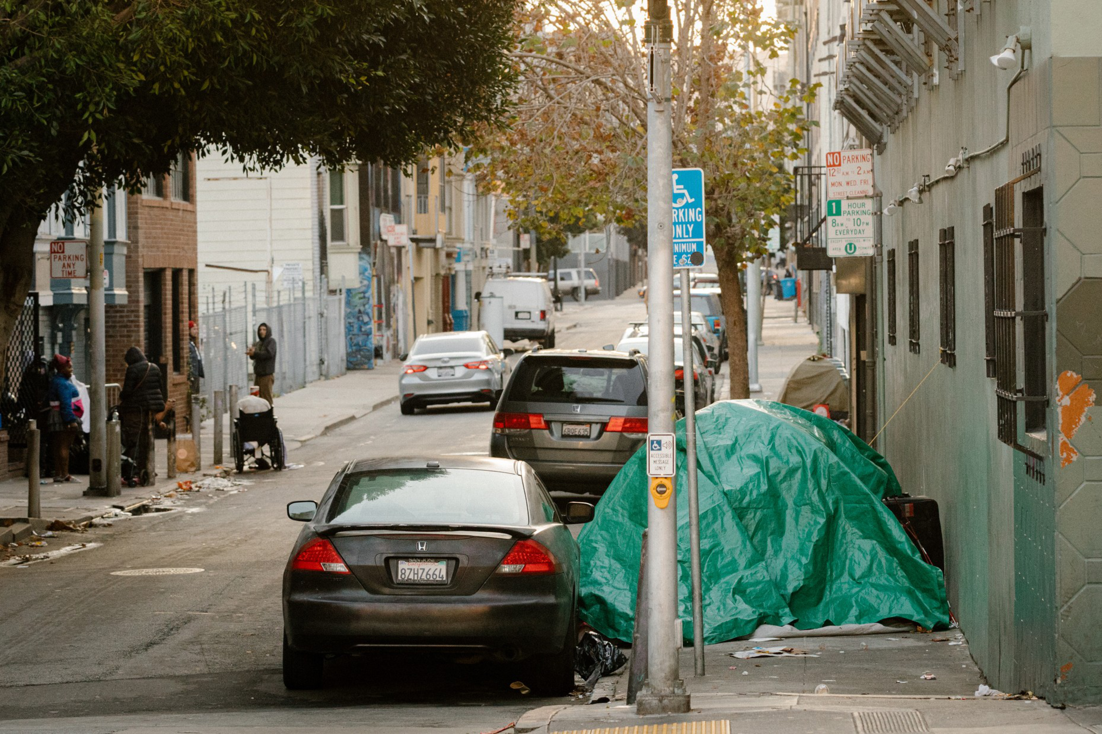 A city street with parked cars, a green tarp-covered tent on the sidewalk, and people standing or walking near buildings littered with trash.