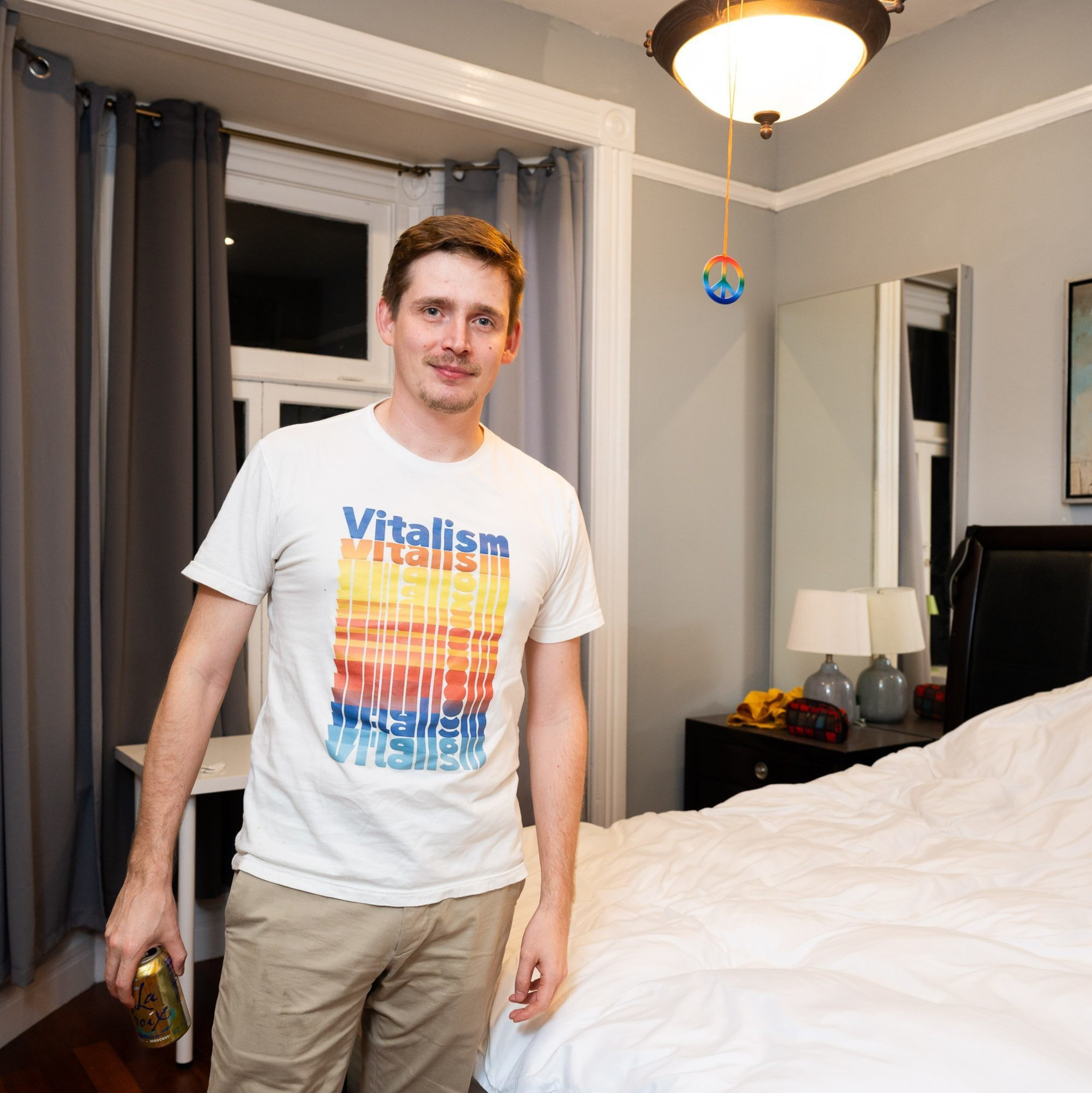 A man smiles in a bedroom, wearing a white &quot;Vitalism&quot; T-shirt and holding a LaCroix can. The room has gray curtains, a bed, and a lamp on a bedside table.