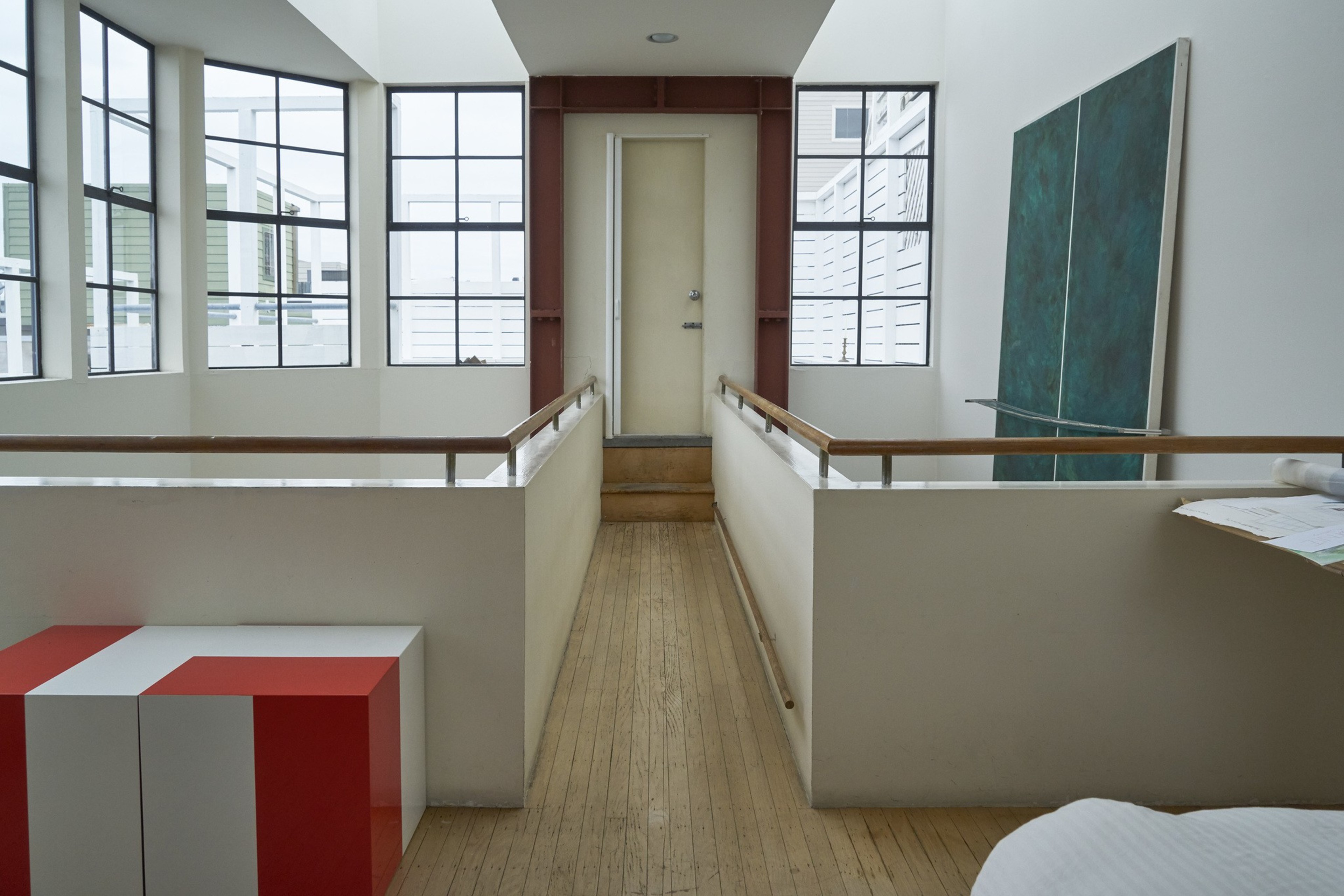 The image shows a modern hallway with light wood flooring, a door at the end, large windows on both sides, a red and white bench, and a large teal artwork.